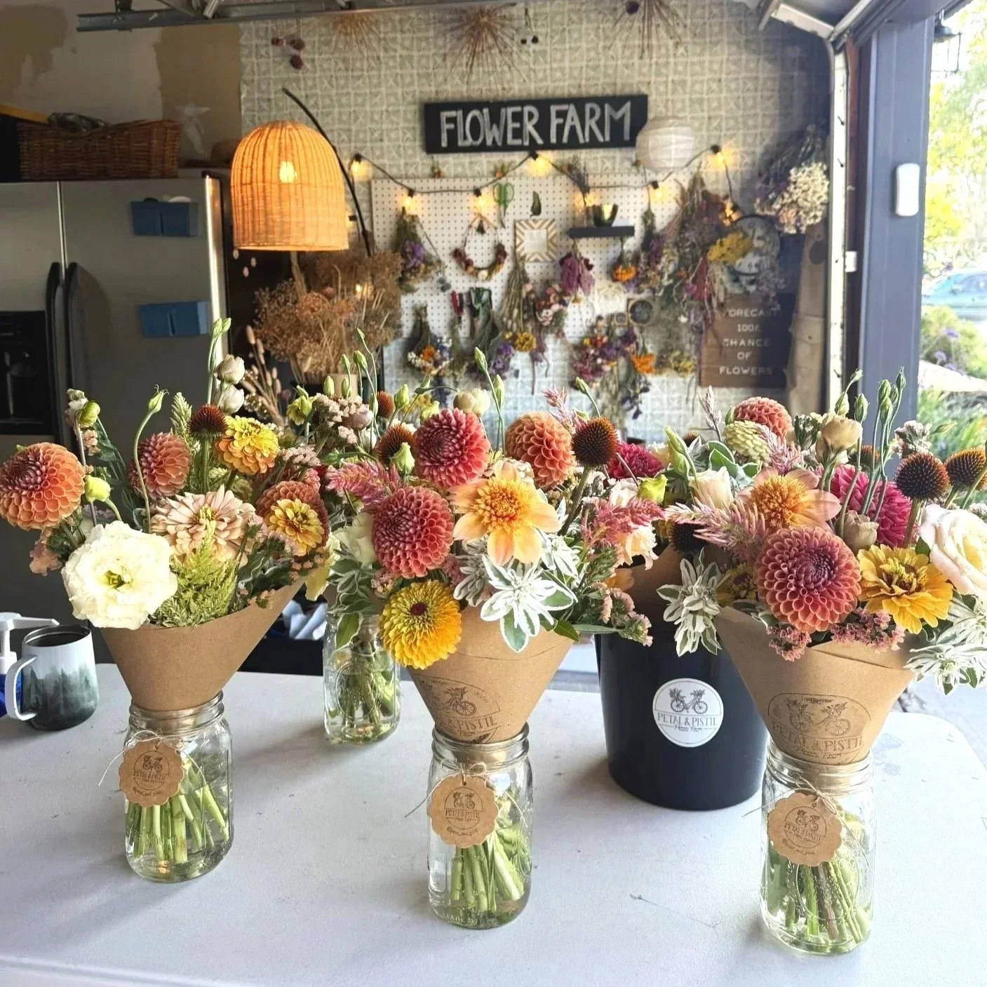 Three mason jars with bouquets of colorful flowers on a table in front of a flower farm workshop area.