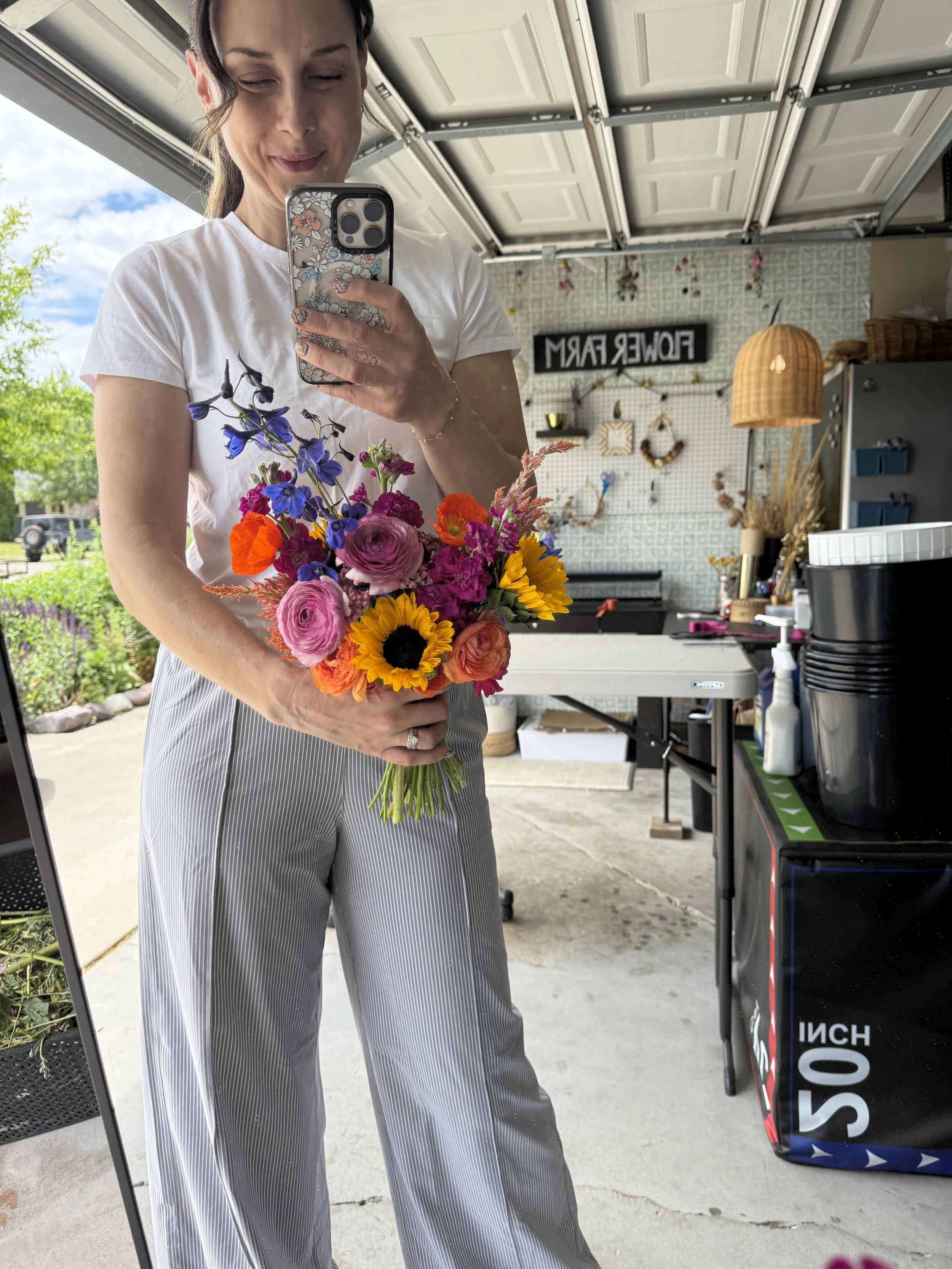 A woman taking a selfie with a bouquet of colorful flowers in front of her. She is standing in a garage with a row of closed garage doors and various decorations on the wall behind her.