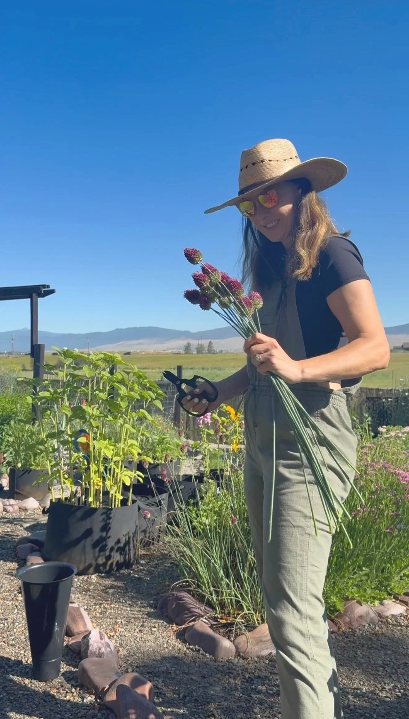 A woman wearing a wide-brimmed straw hat, sunglasses, and khaki pants is holding a bunch of flowers and gardening shears, standing in a garden with various plants and flowers under a clear blue sky.