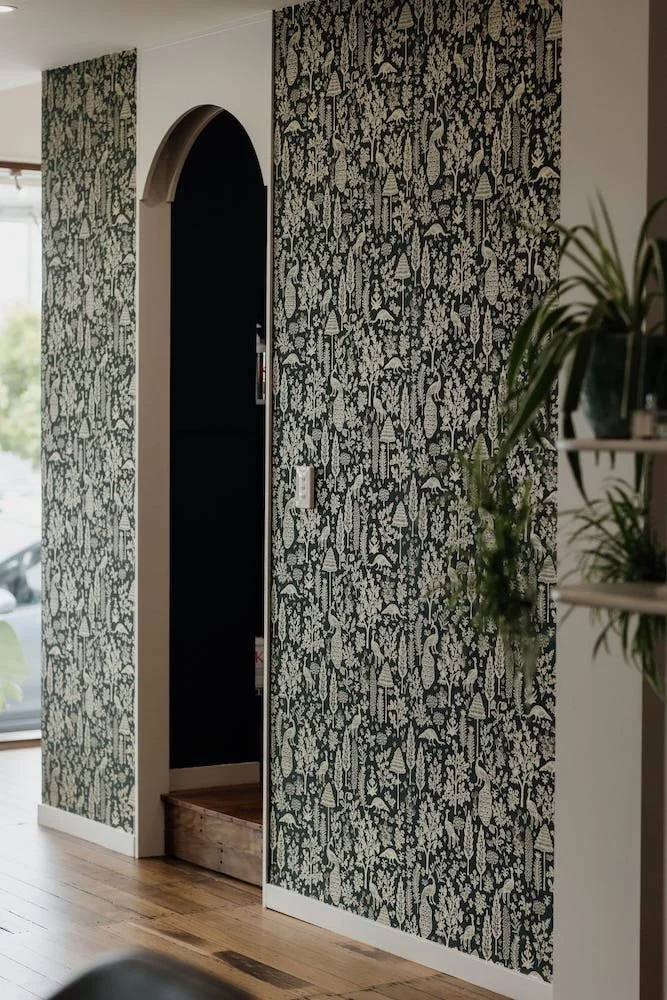 Interior of a room with floral-patterned wallpaper in black and white, an arched doorway leading to a dark room, and a wooden platform at the base of the doorway. A plant with long green leaves is partially visible on the right.