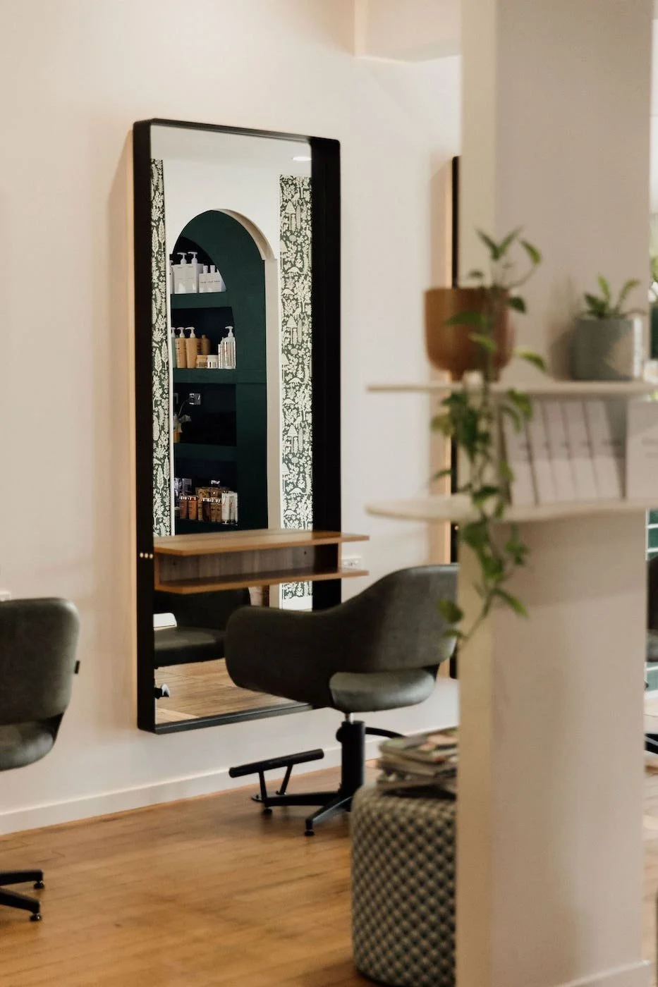A salon or hair studio with a large mirror framed in black, reflecting shelves with hair products, and a styling chair with a small desk beneath the mirror. There are some plants and a white partition in the foreground.