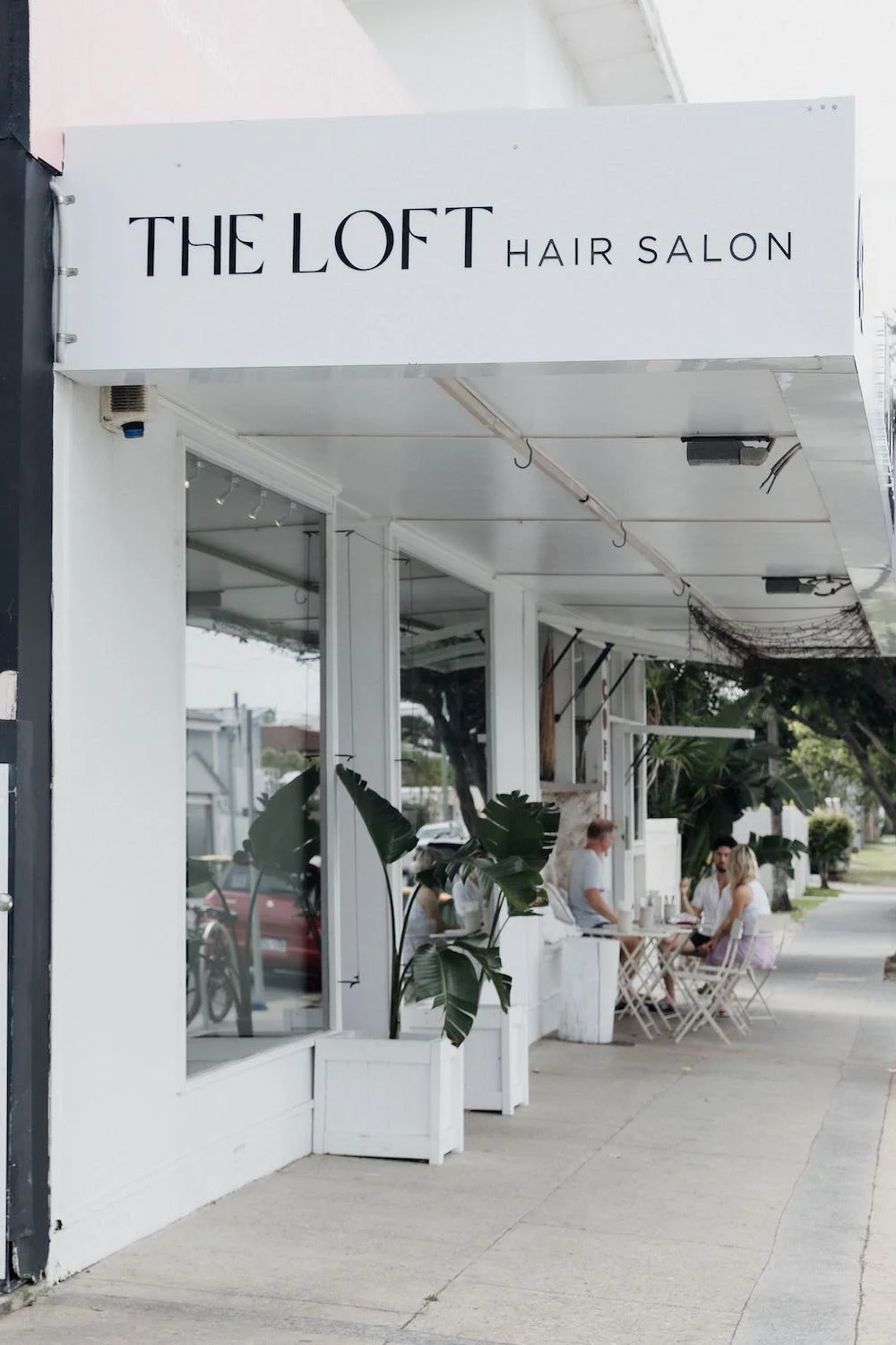Exterior of The Loft Hair Salon with white facade, large windows, and outdoor seating with people, potted plants, and trees in the background.