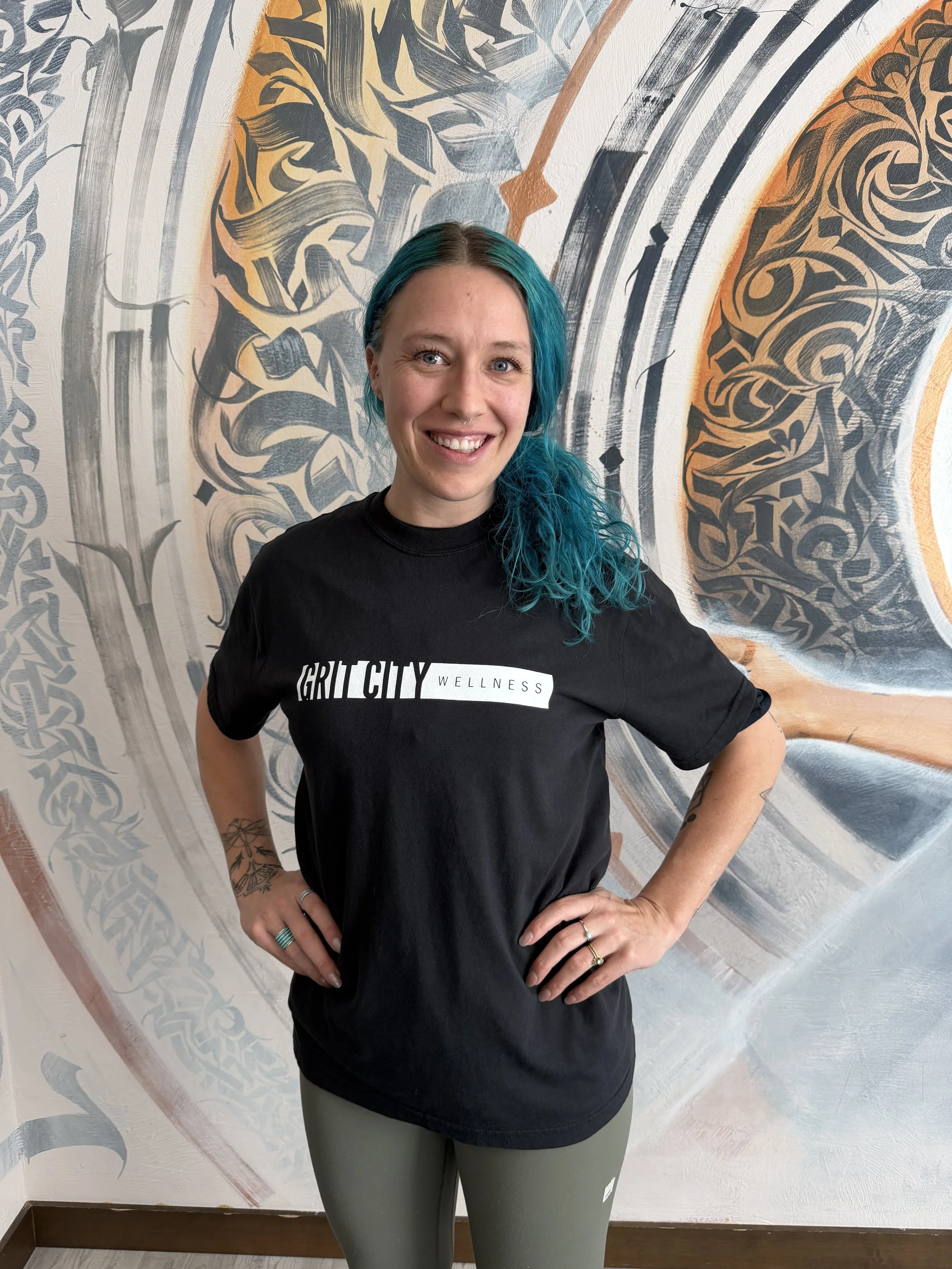 A smiling woman with long, curly blonde hair wearing a black shirt in front of a mural with intricate black, gray, orange, and white designs.