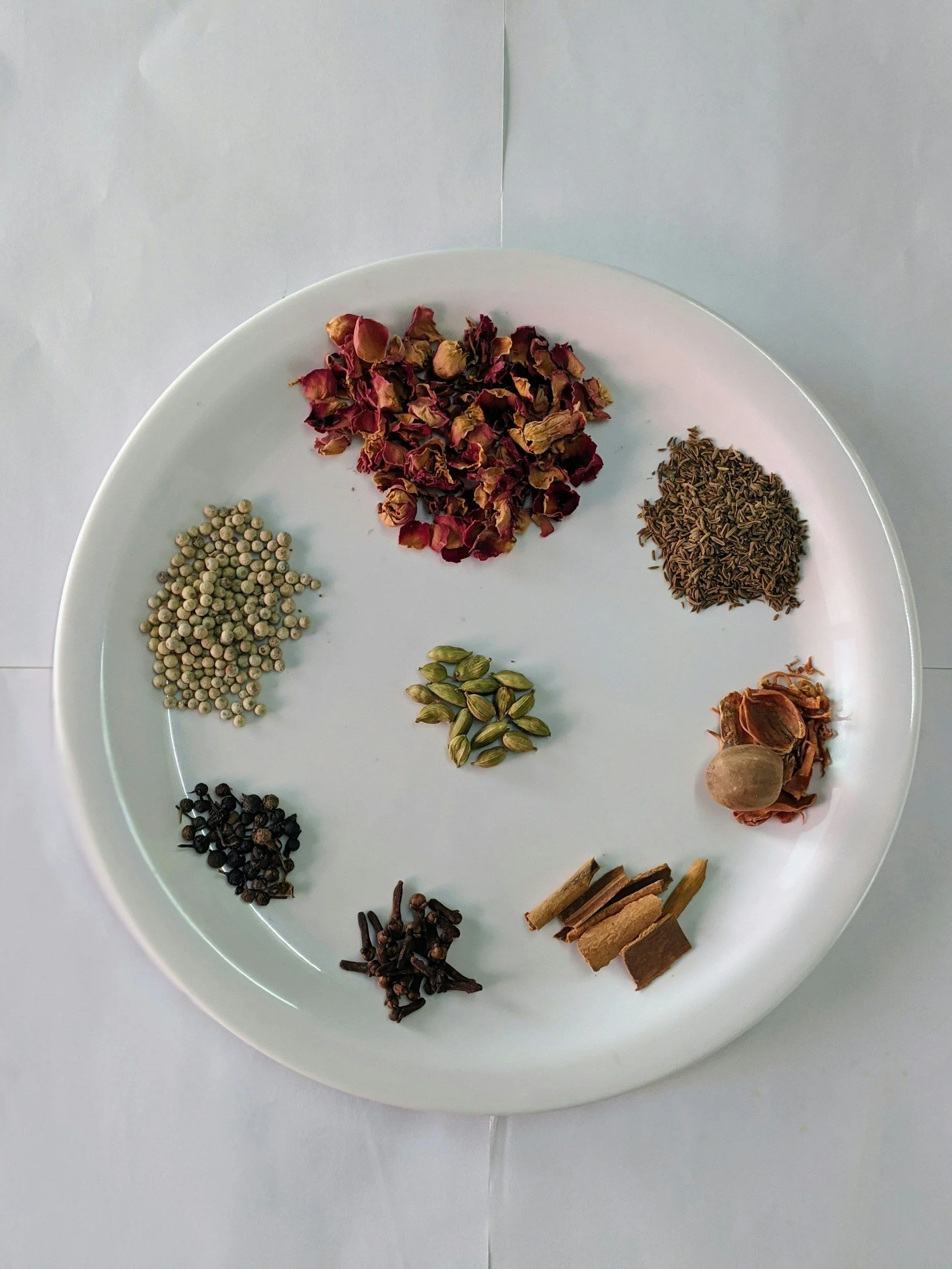 A white plate containing various spices and herbs, arranged in a circle, including dried rose petals, cumin seeds, coriander seeds, black peppercorns, cloves, cinnamon sticks, nutmeg, and other dried herbs.