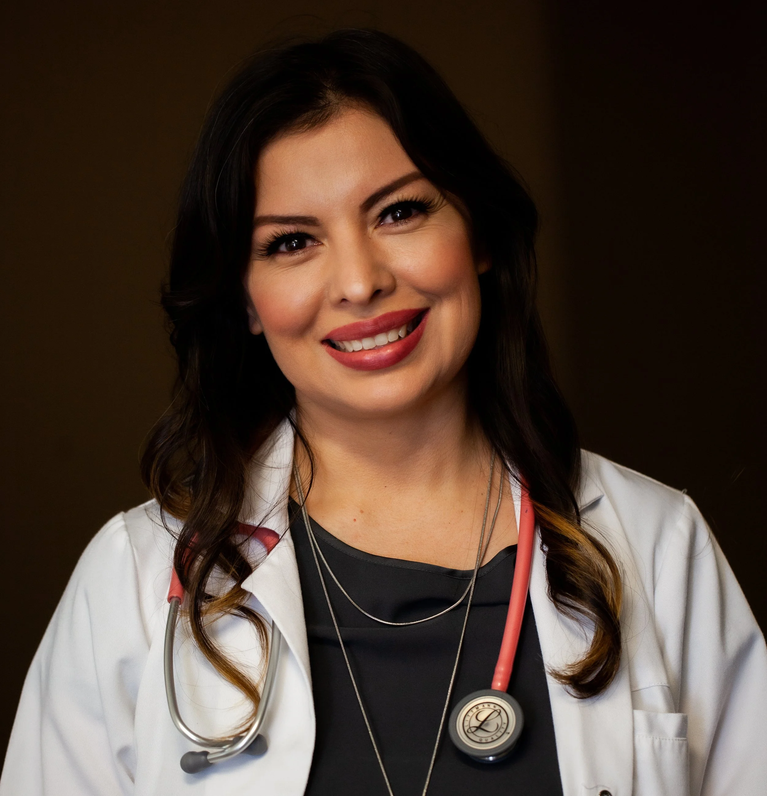Close-up of a smiling female doctor with dark hair, wearing a white lab coat and a stethoscope around her neck, against a dark background.