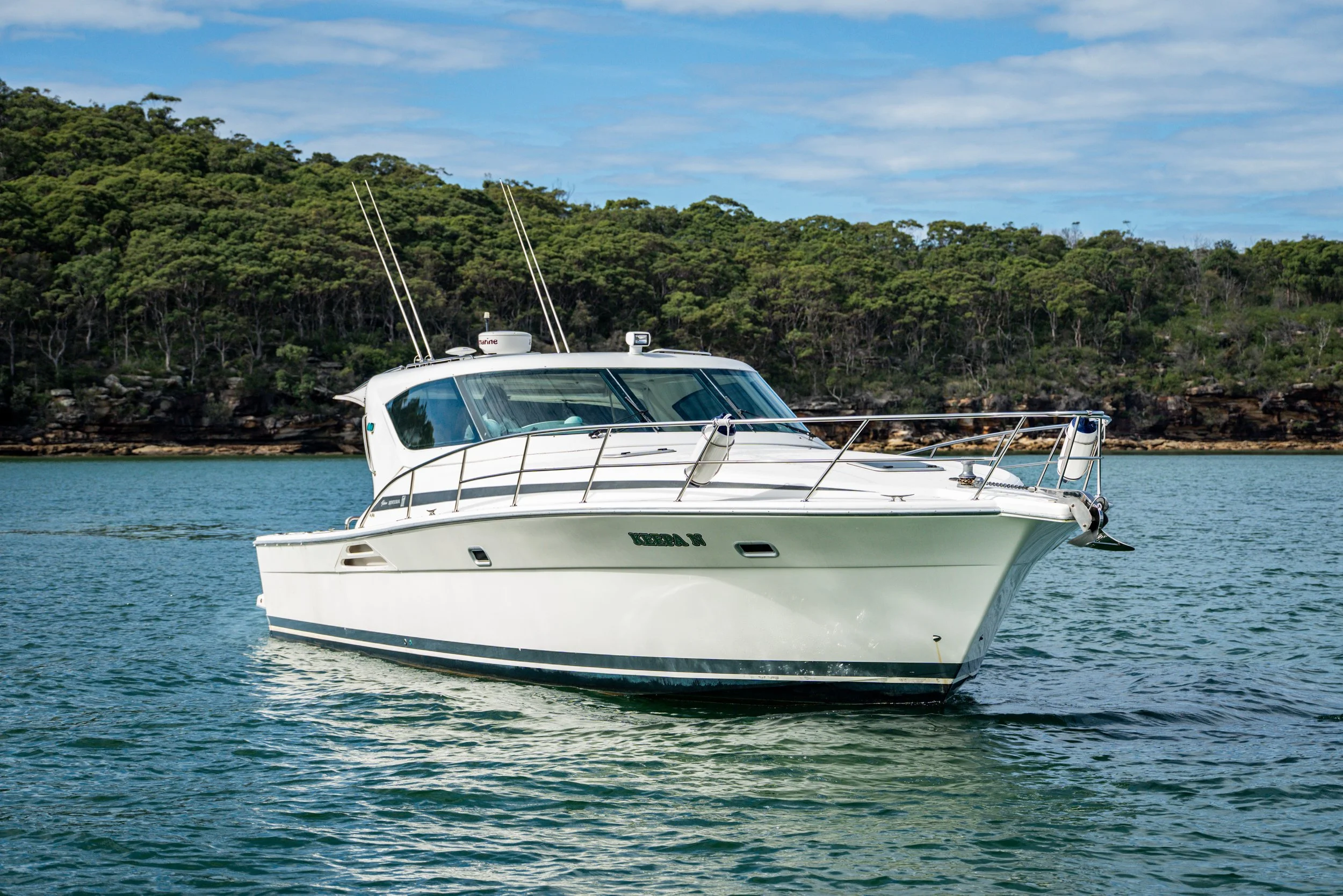 A white motor yacht sailing on calm water with a wooded shoreline and blue sky in the background.