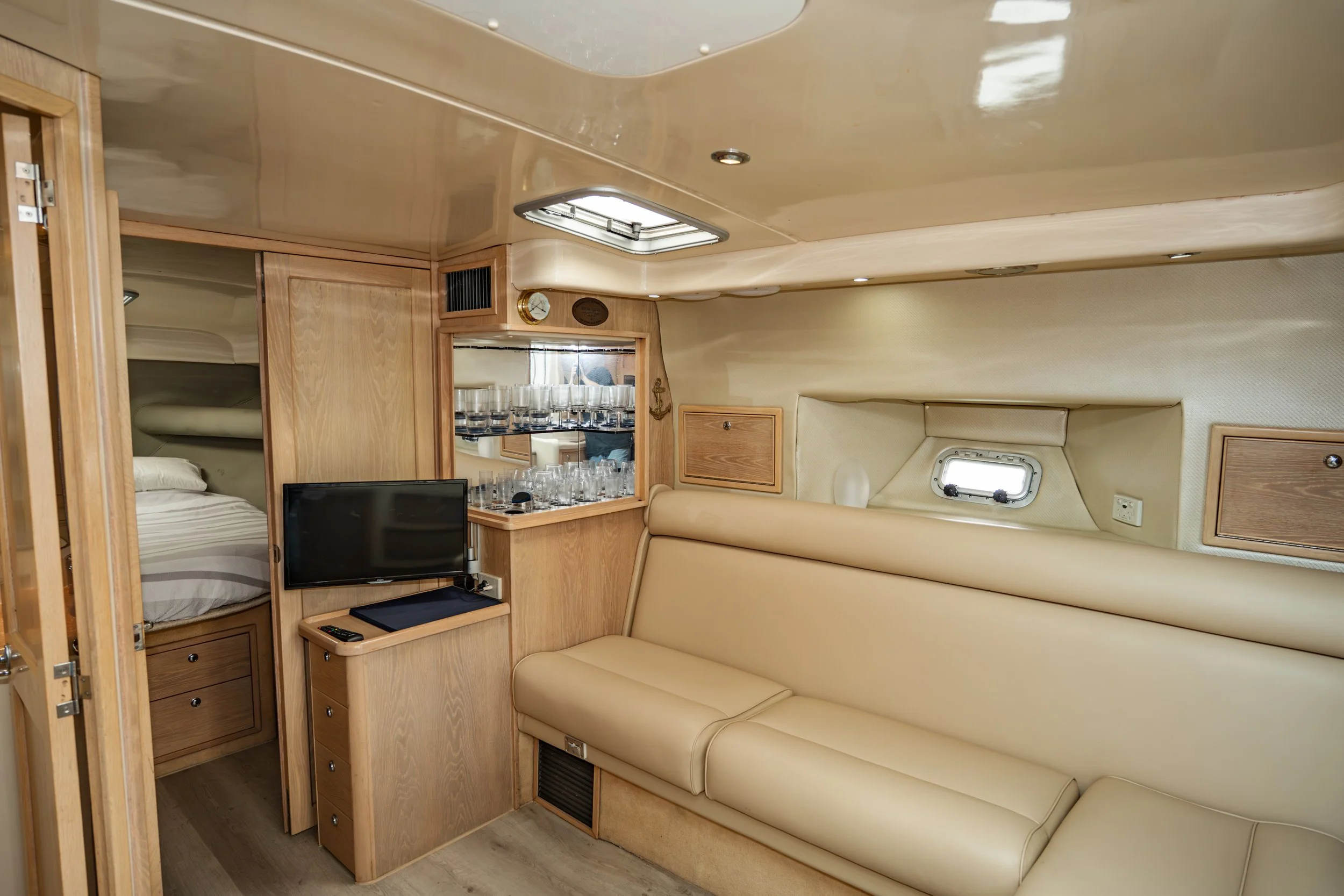Interior of a yacht cabin with a beige leather sofa, a small flat-screen TV, a wooden cabinet with glassware, and a bed with a sliding door, wood paneling, and small windows.