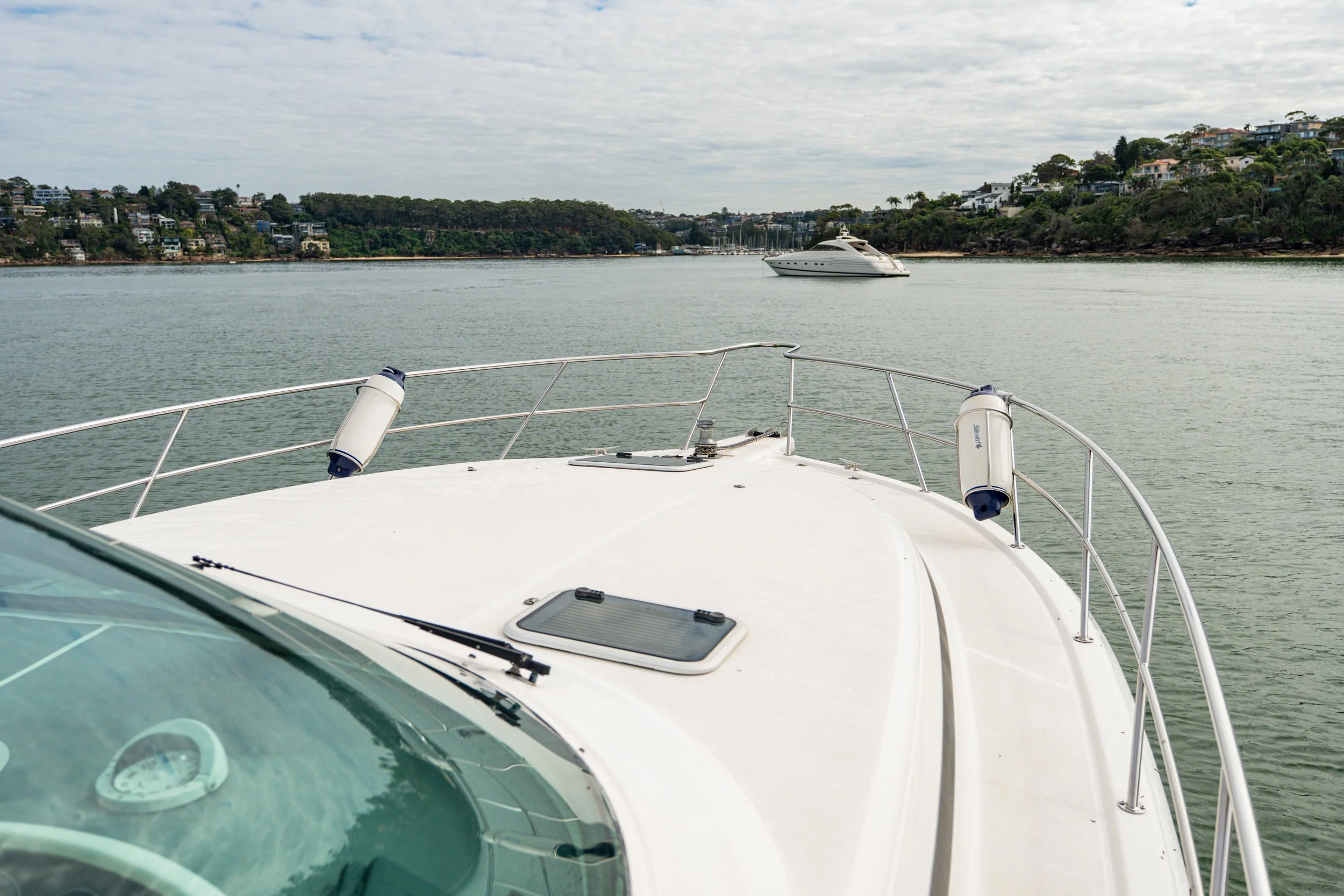 View from the bow of a yacht looking out over calm water, with another yacht anchored in the distance and residential houses on green hills along the shoreline under cloudy skies.
