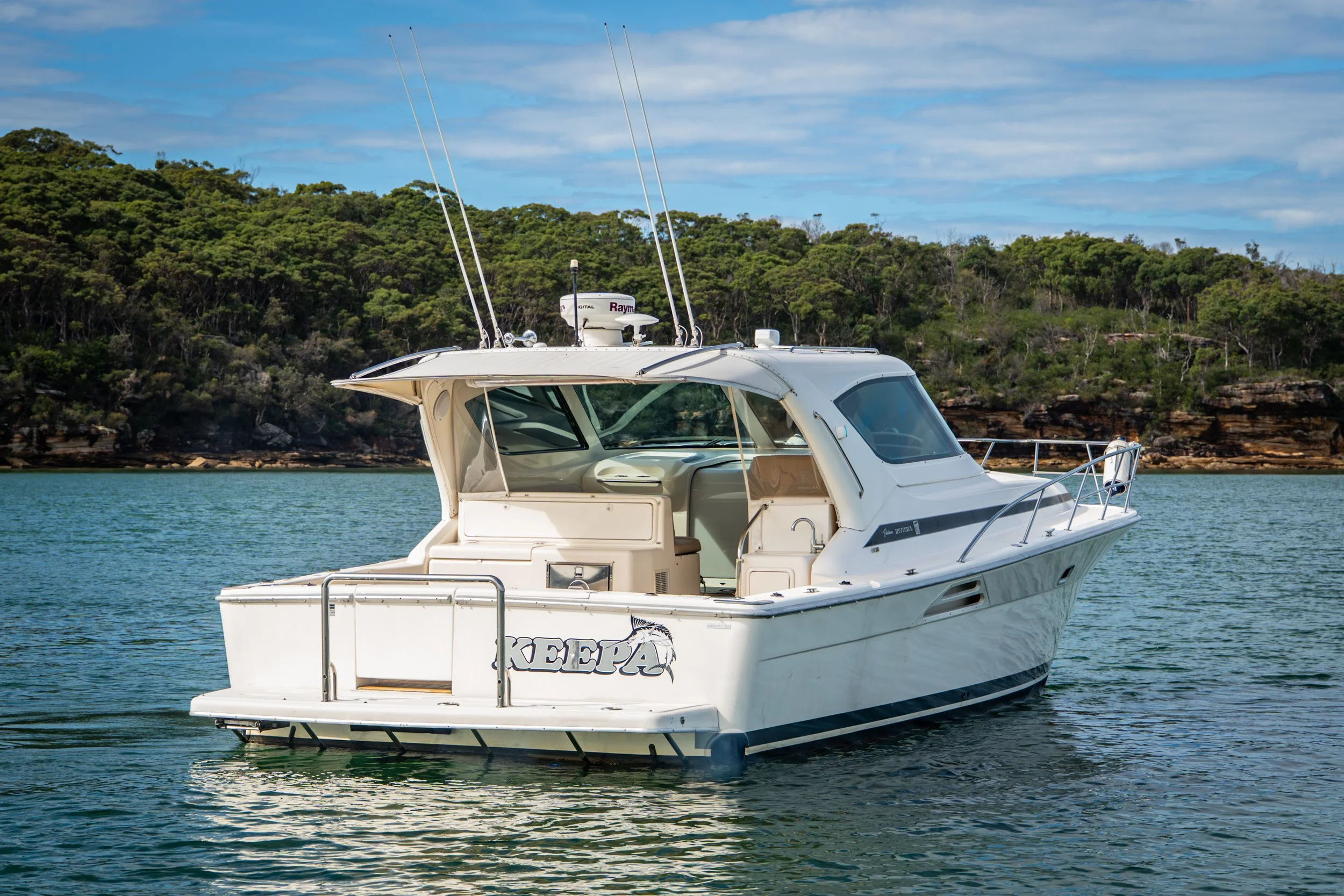 A white motor yacht named KEETAPA floating on calm water, with a lush green hilly shoreline in the background and a partly cloudy sky overhead.