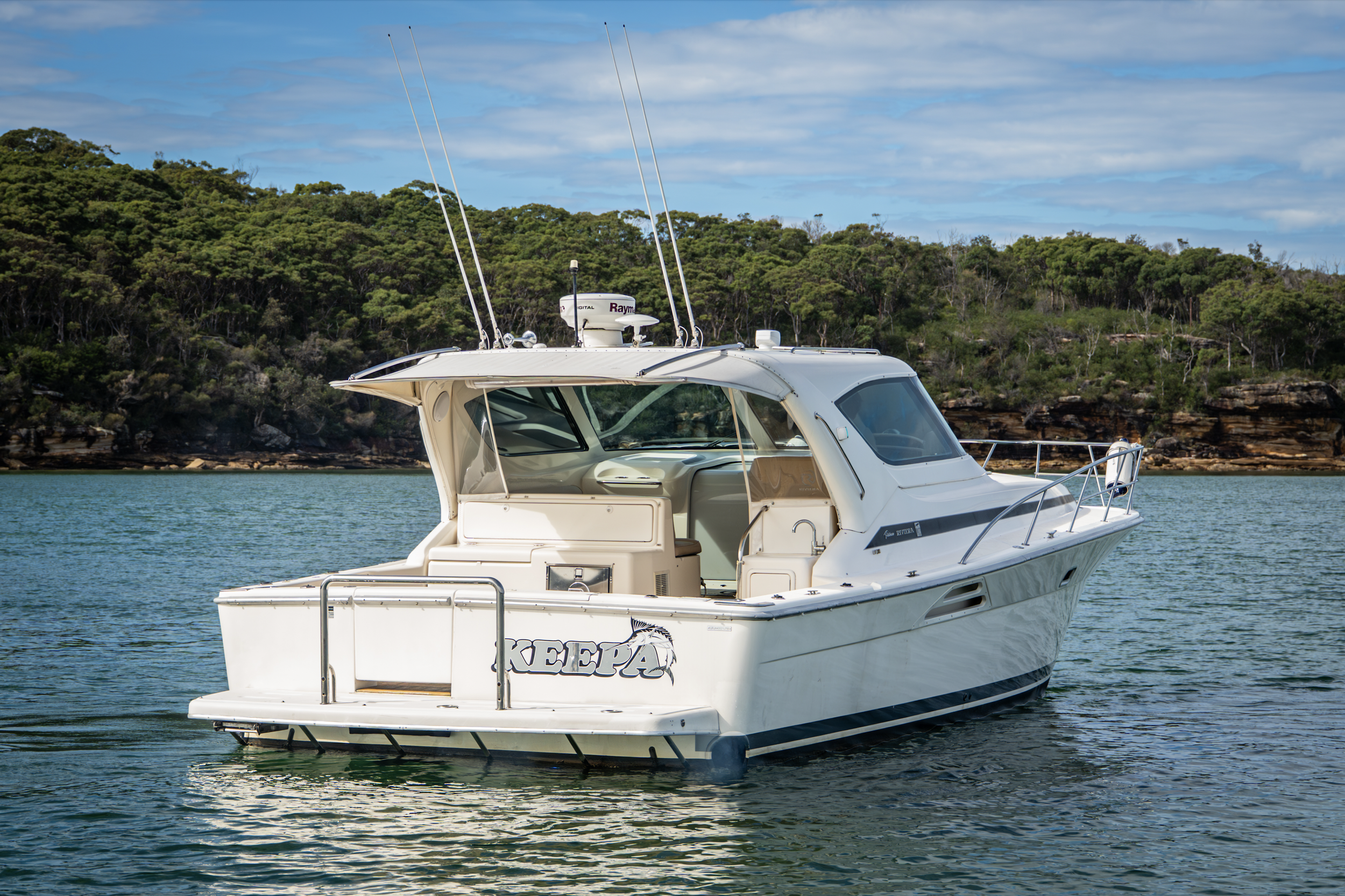 A white motor yacht named 'KEEPA' floating on water near a green, forested shoreline with a partly cloudy sky overhead.