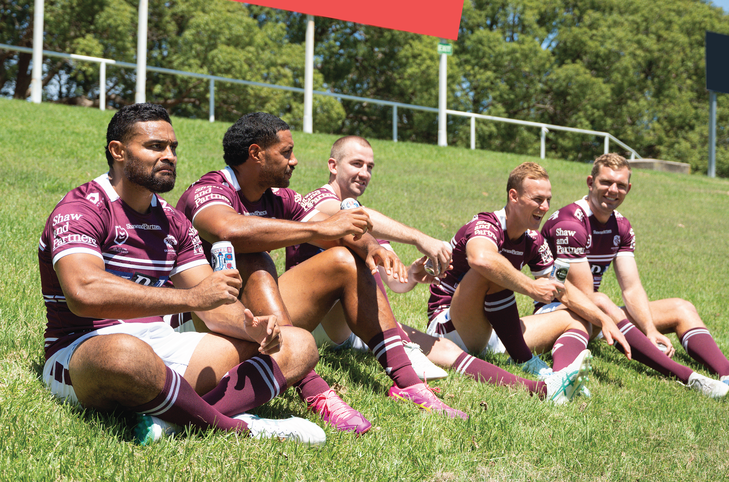 Group of five rugby players sitting on grass field, wearing matching maroon jerseys and shorts, enjoying drinks during a sunny day.