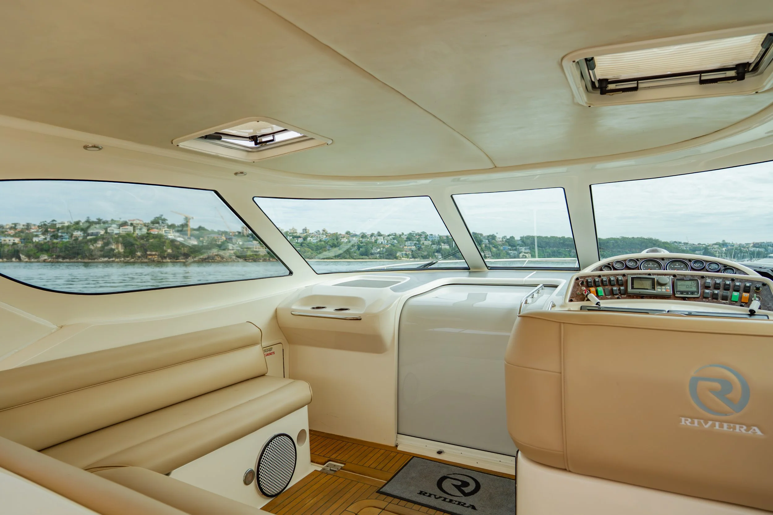 Interior of a luxury boat with beige leather seating and navigation controls, overlooking water and shoreline with houses.