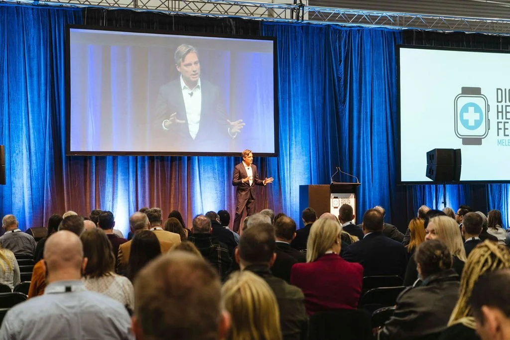 A speaker on stage at the Digital Health Festival addressing an audience, with large screens displaying his presentation.