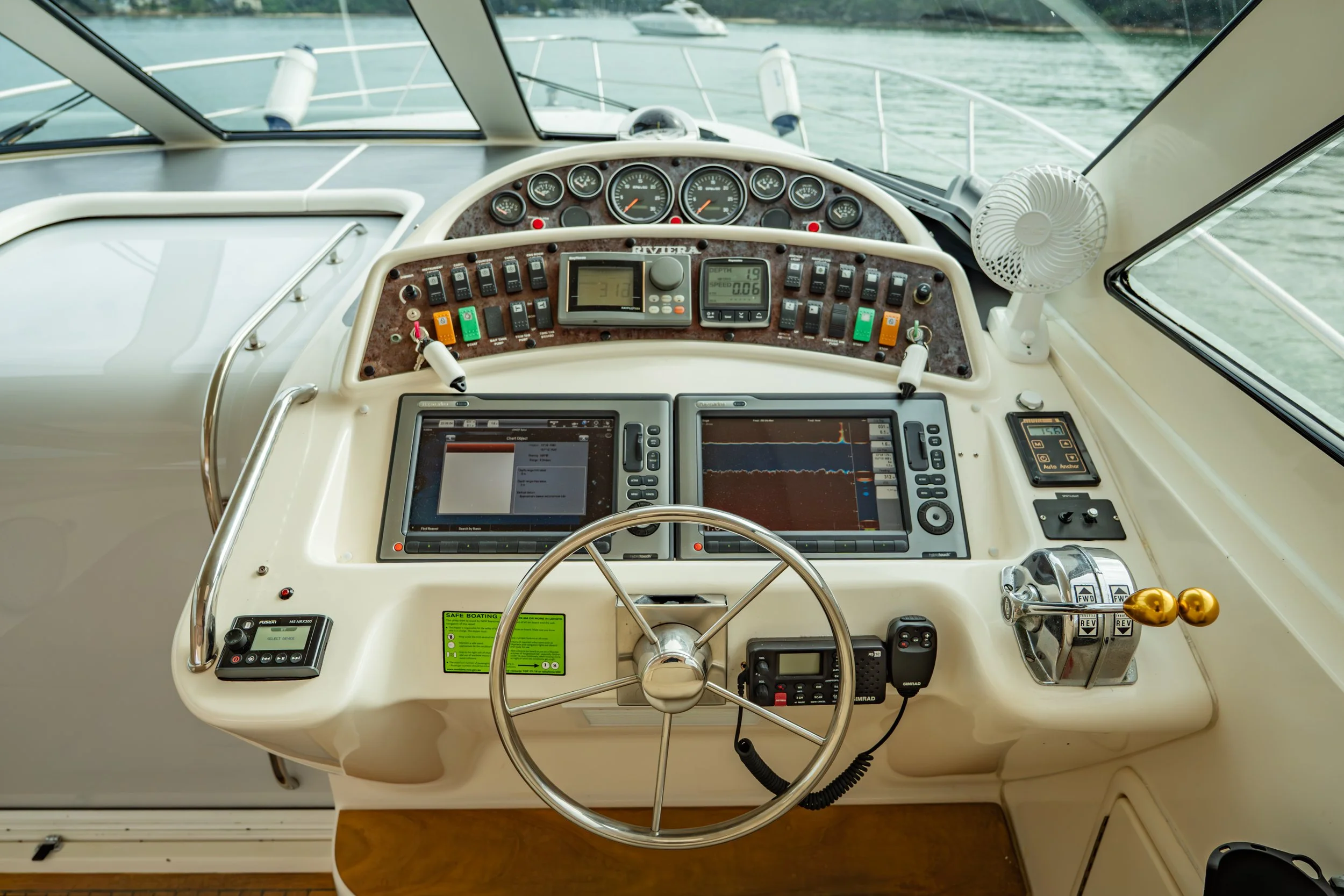 View of a yacht's cockpit with steering wheel, electronic navigation screens, throttle controls, and instrument gauges, with water visible through the windows.