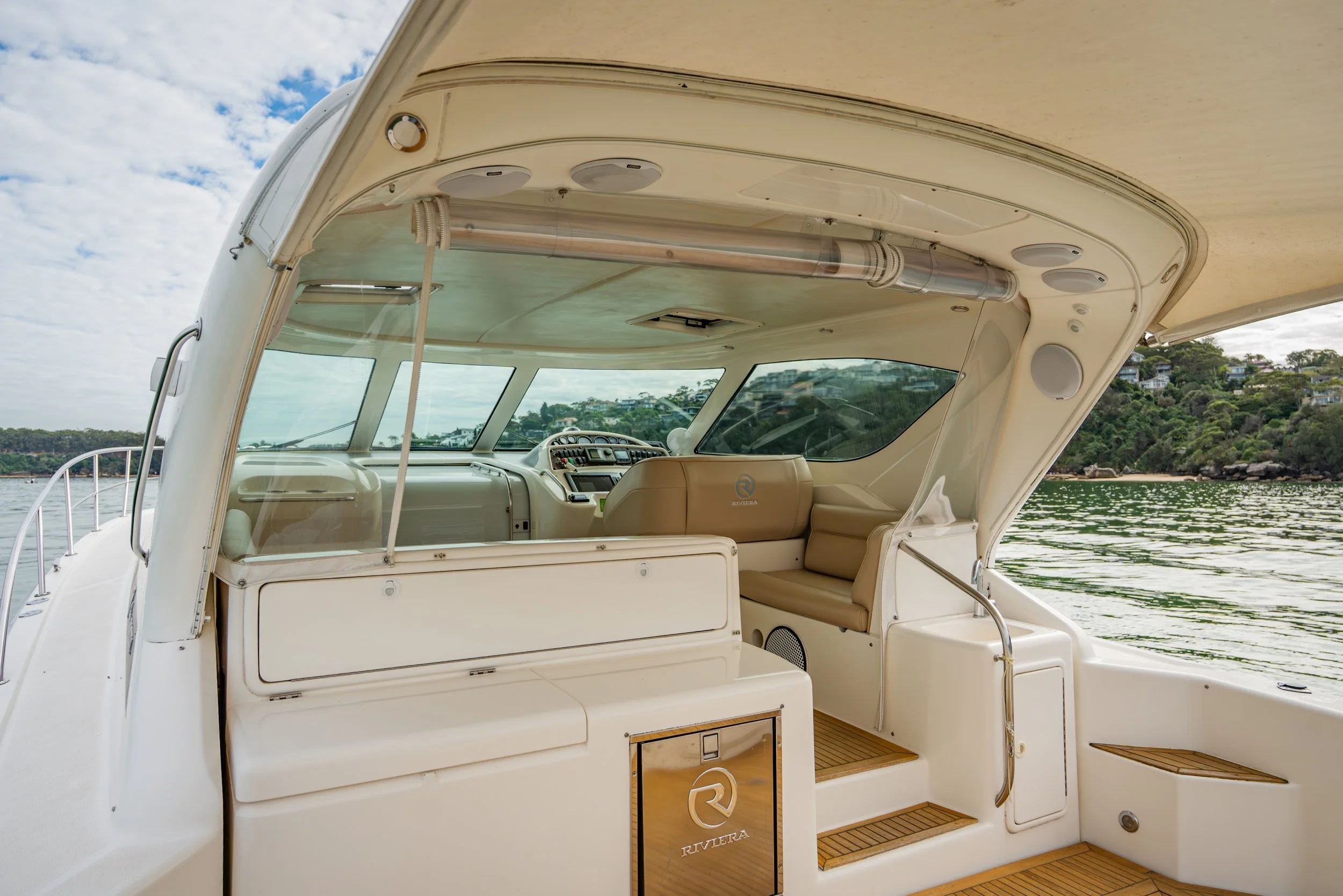 Interior of a luxury boat showing beige leather seats, control panel, large windows, and a view of water and coastline in the background.