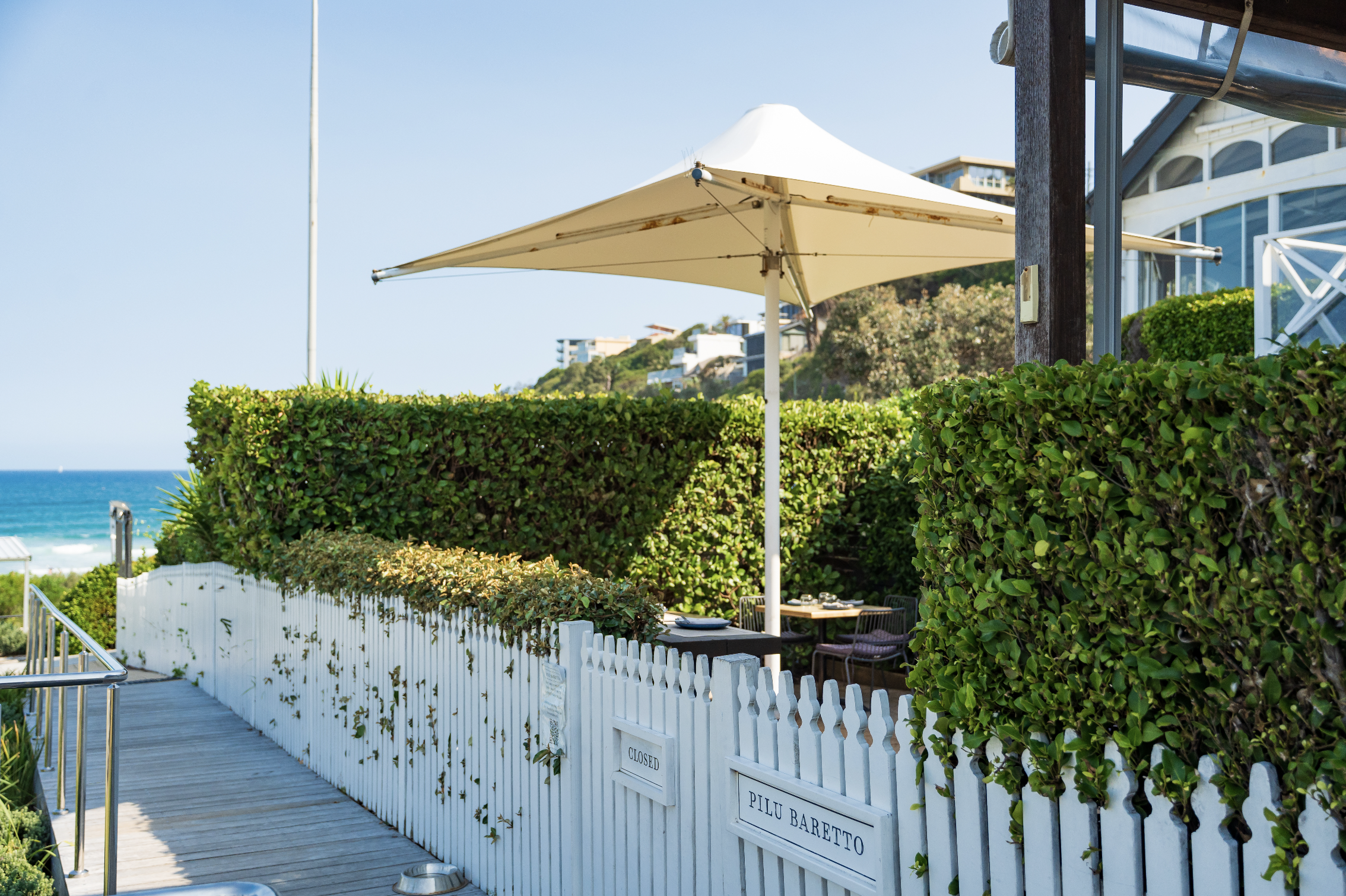 Outdoor seating area with a white fence, green bushes, an umbrella, and views of the ocean and buildings in the background.