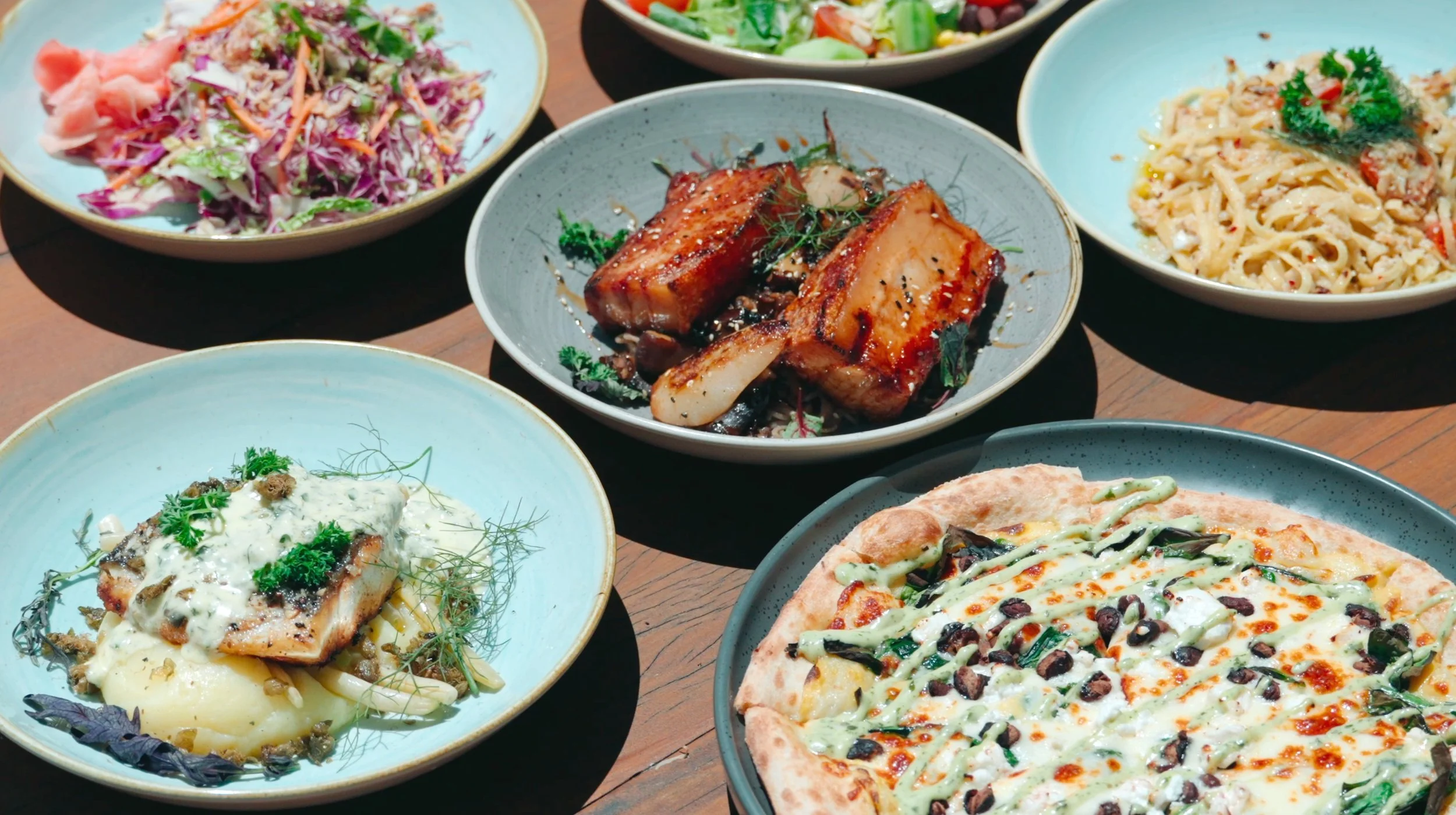 Assorted dishes including salad, grilled ribs, pasta, fish, and pizza on a wooden table