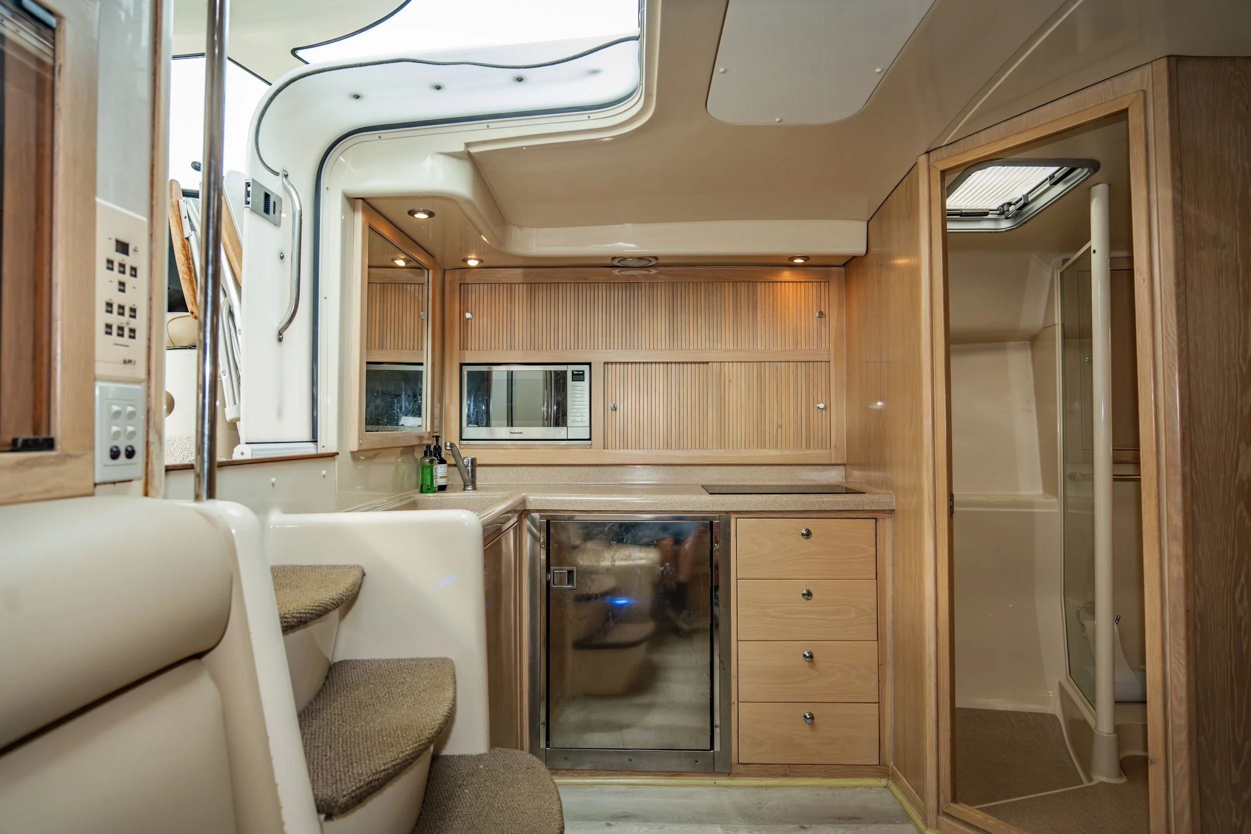 Interior of a small boat or RV kitchen with light wood cabinets, a microwave, a sink, and a small refrigerator, with a partially visible staircase and a shower enclosure.