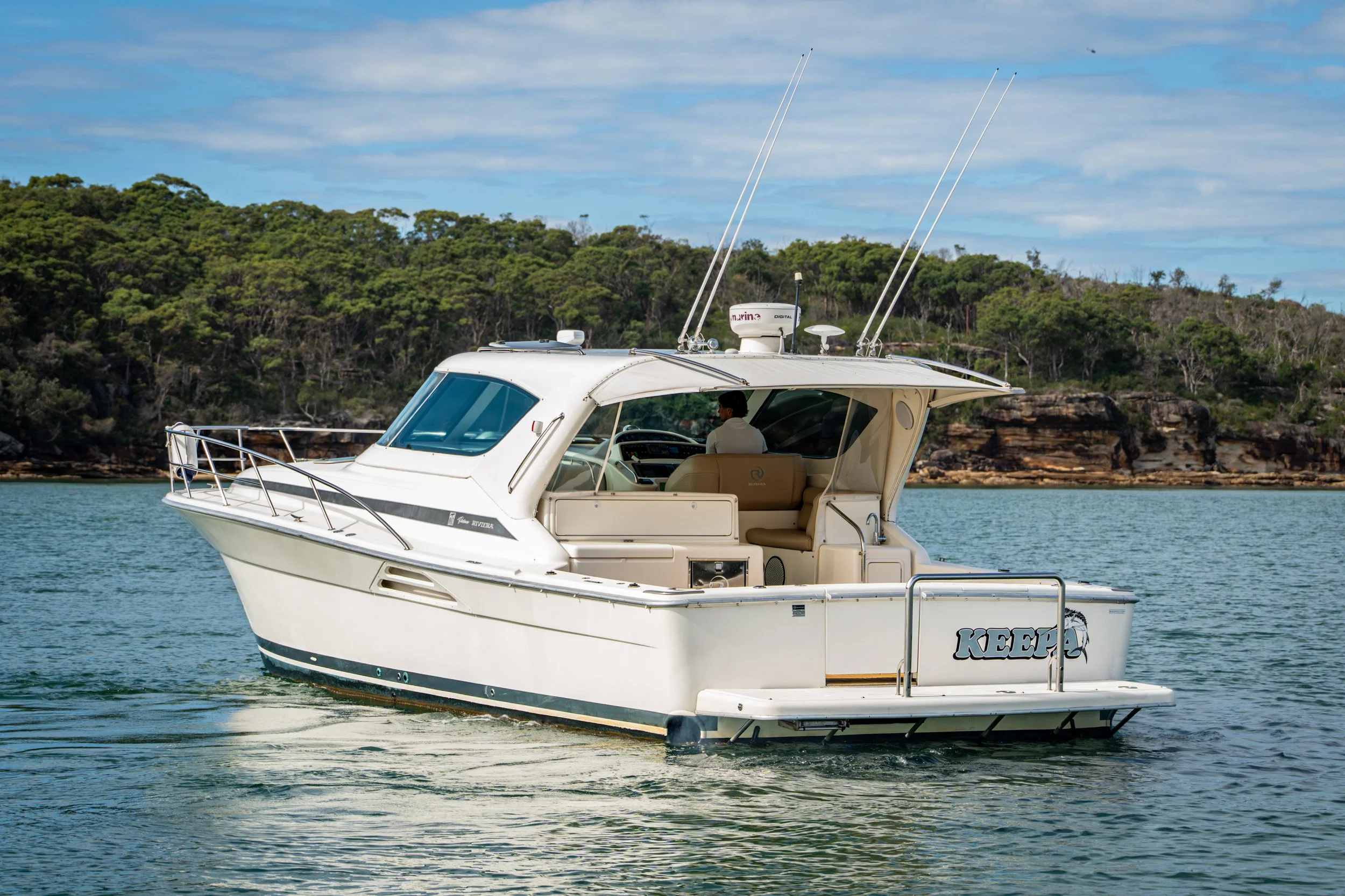 White motor yacht named 'KEEPER' cruising on calm water with green forested shoreline in the background.