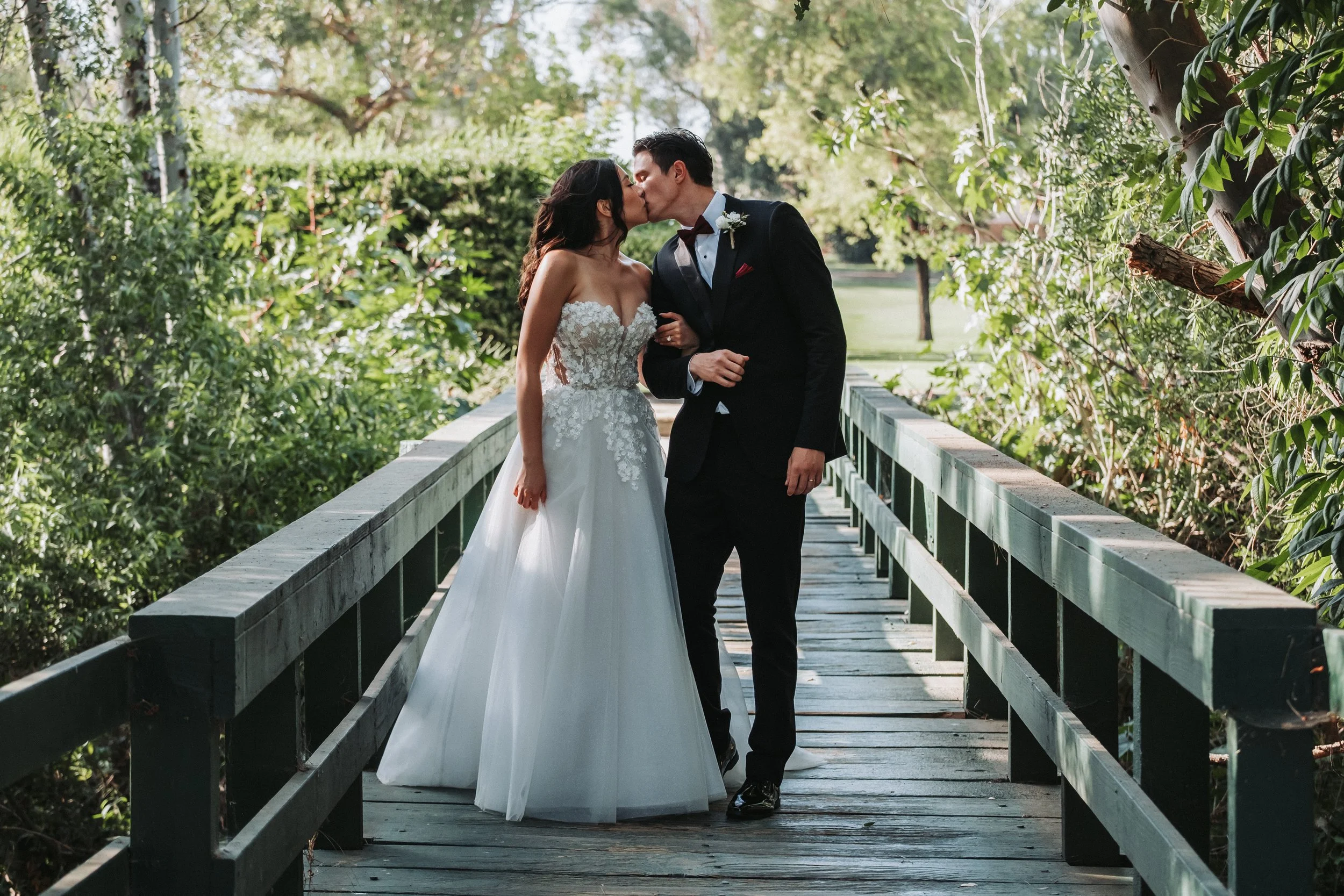 A bride and groom kiss on a wooden bridge surrounded by green trees and bushes.