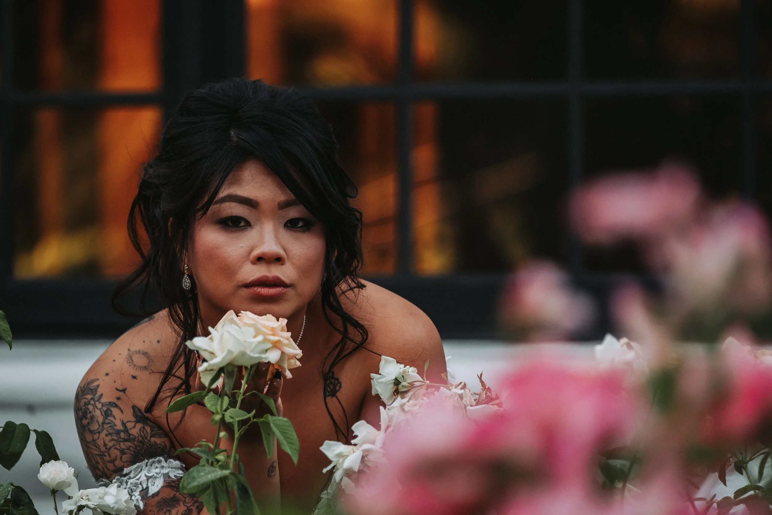 A woman with dark hair and tattoos on her shoulders and arms, surrounded by pink and white flowers, with a black grid structure and warm lighting in the background.