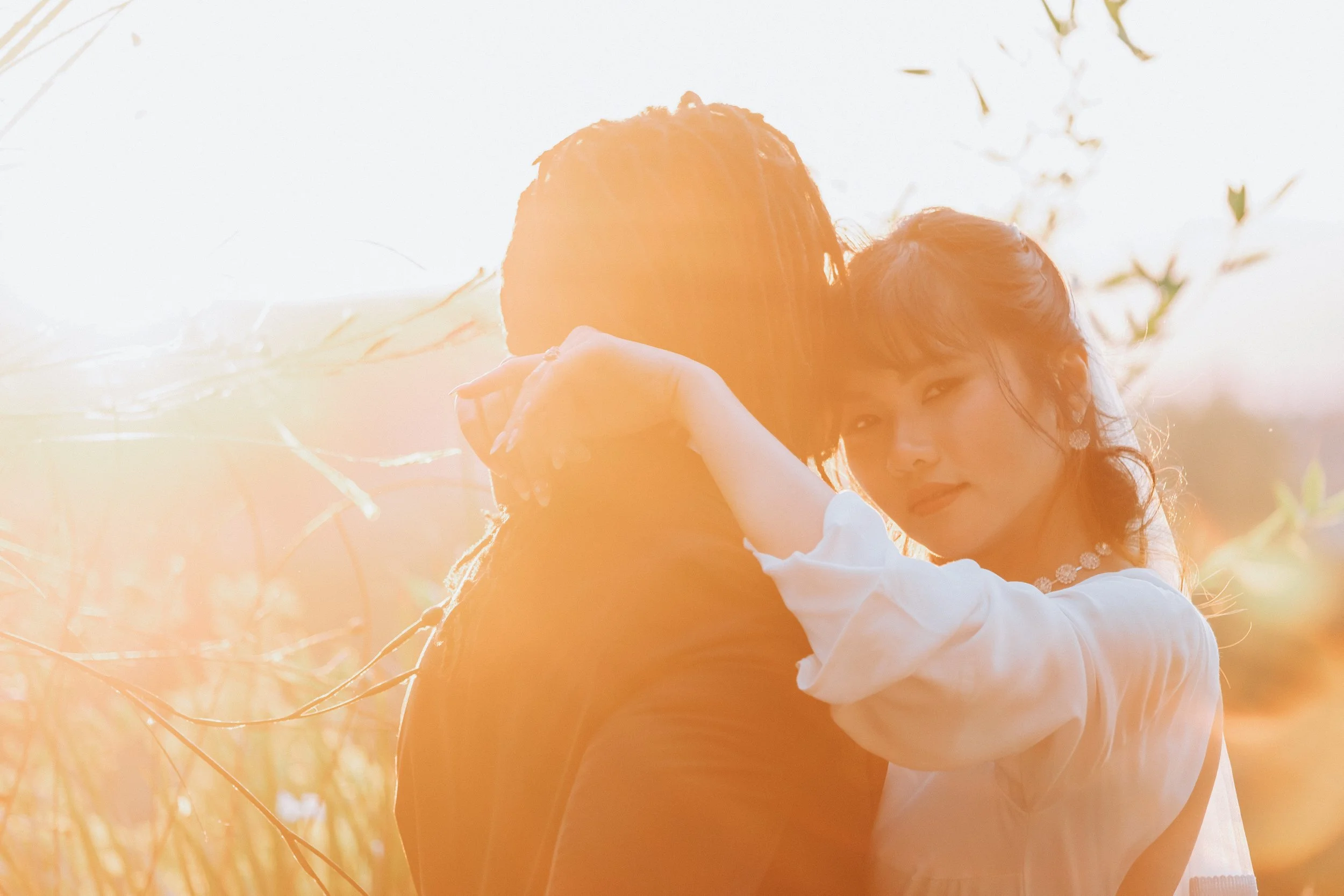 A woman with dark hair, wearing jewelry and a light-colored dress, hugs a person with long dark hair from behind in a bright outdoor setting with sunlight filtering through trees.