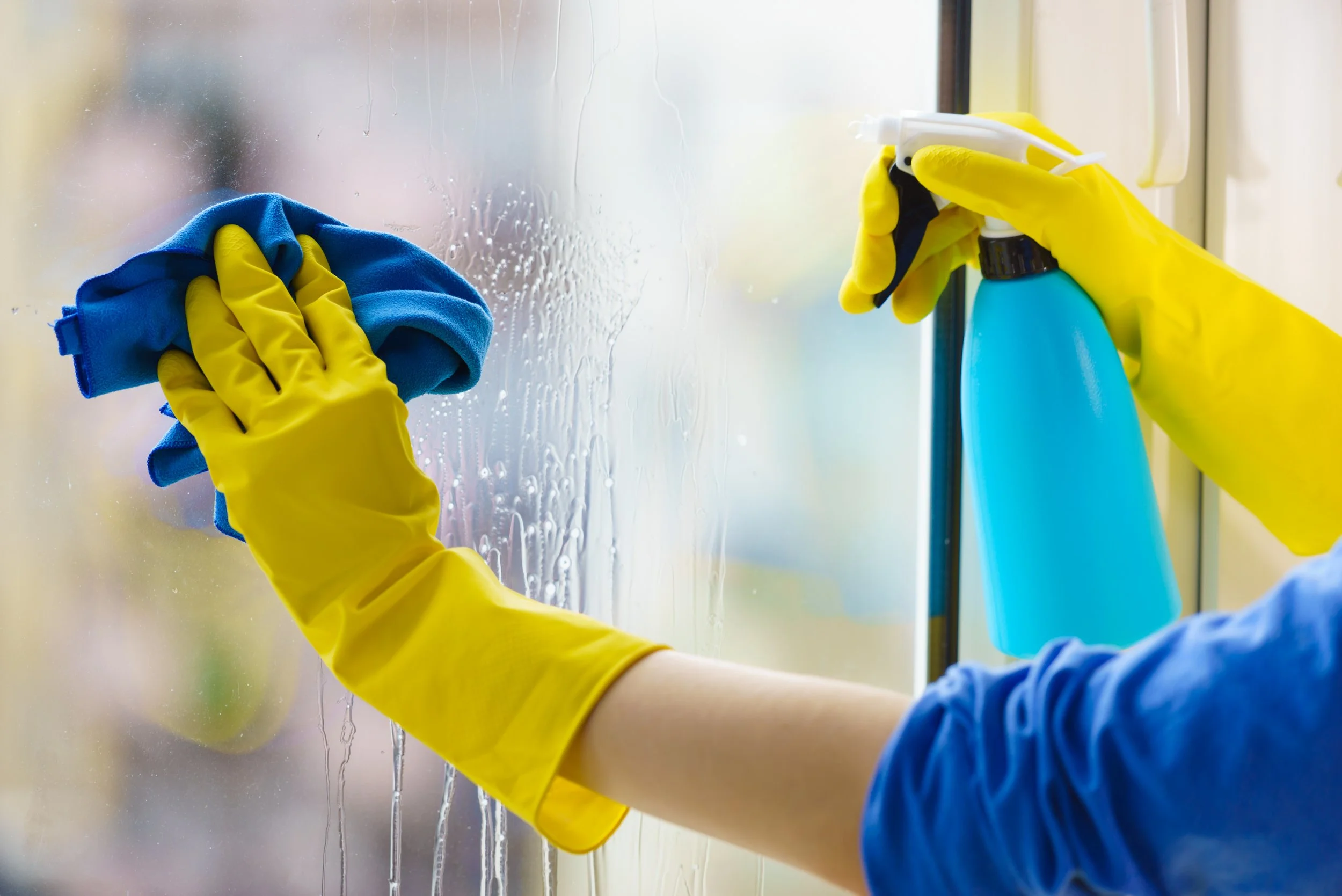 Person wearing yellow rubber gloves cleaning a window with a blue cloth and a spray bottle of cleaning solution.