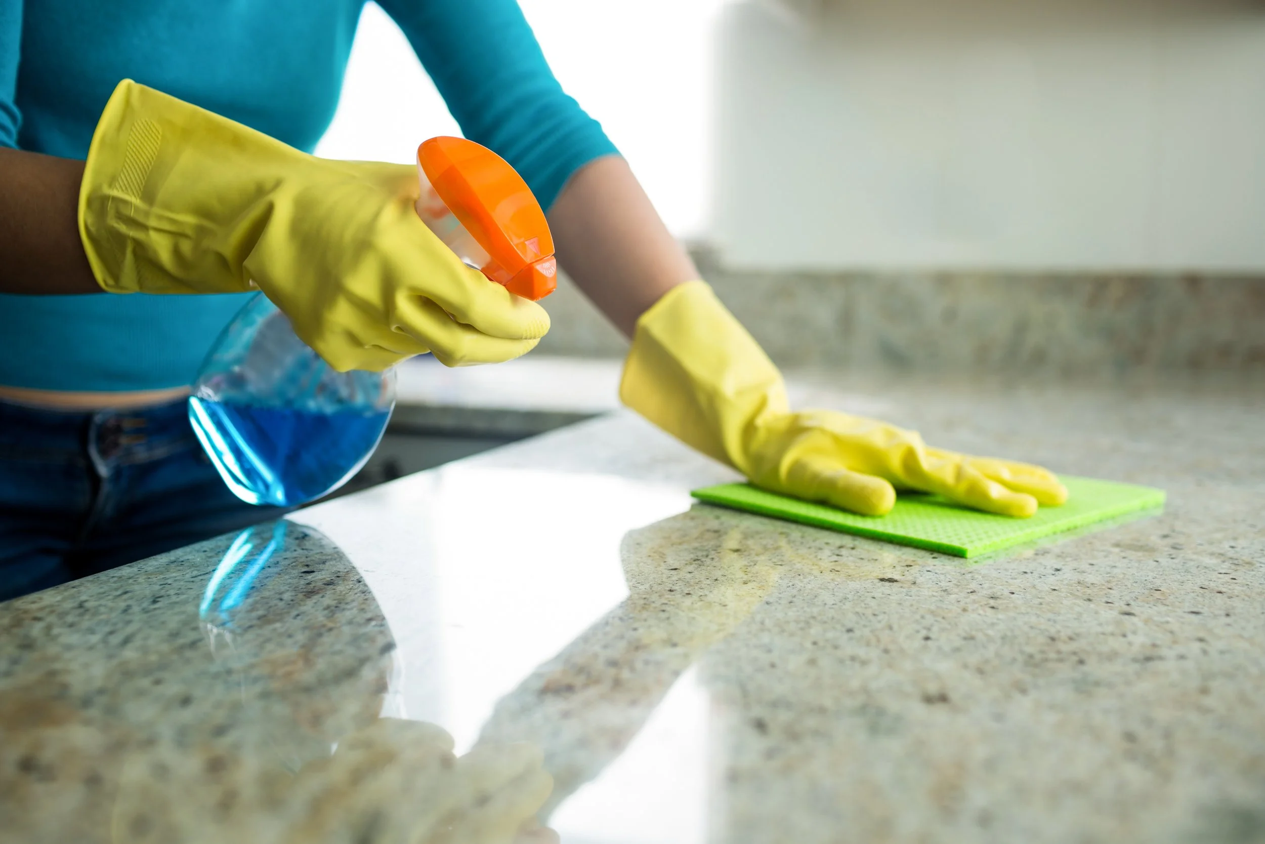 Person cleaning a kitchen countertop with a spray bottle of blue cleaner and wearing yellow rubber gloves.