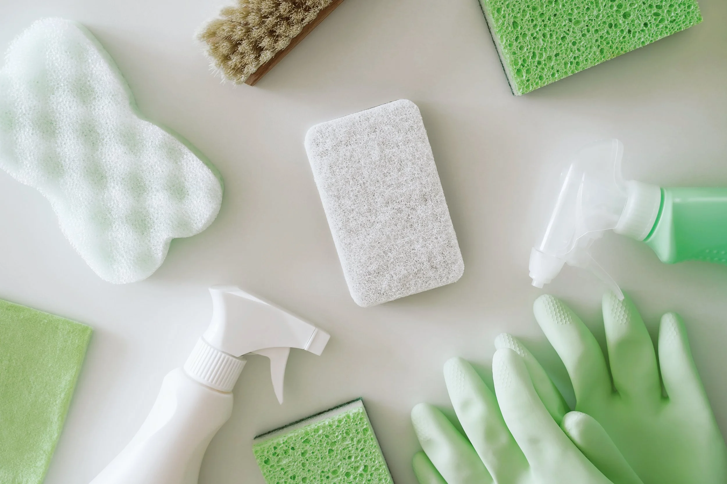 Cleaning supplies including a white spray bottle, a green spray bottle, a pair of green rubber gloves, a white sponge, green and yellow sponges, a scrub brush, and a white cleaning cloth on a white surface.