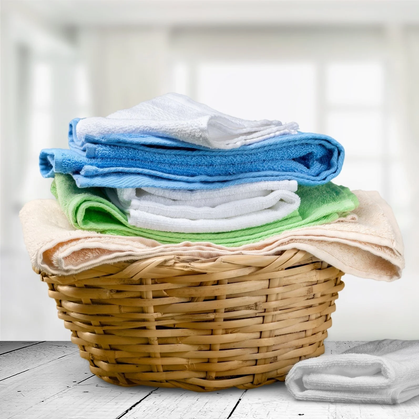 A wicker basket filled with folded colorful towels on a white wooden surface, with a white towel outside the basket.