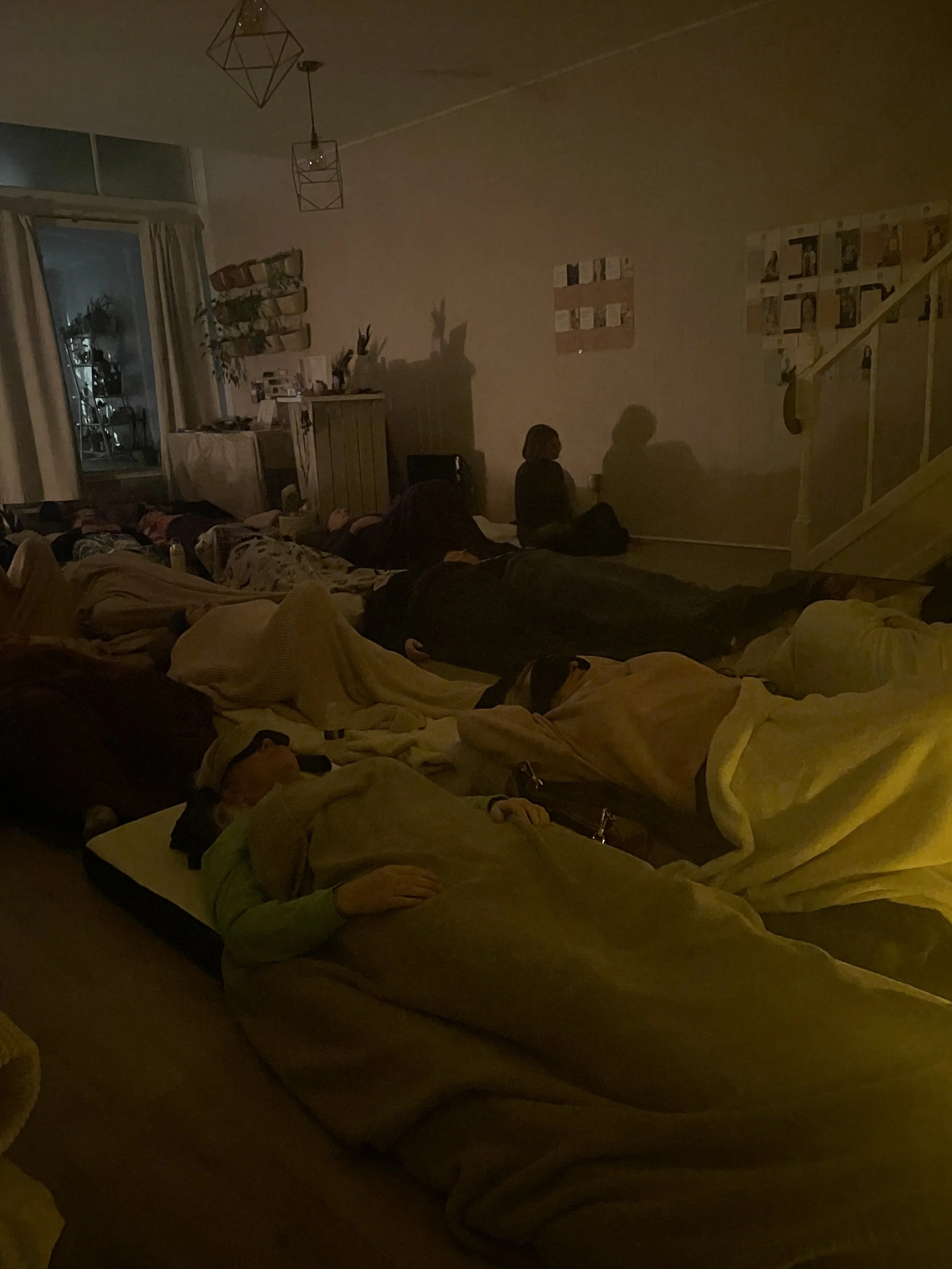 Four people lying on their backs with eyes closed, relaxing on colorful blankets, in a room filled with green houseplants.