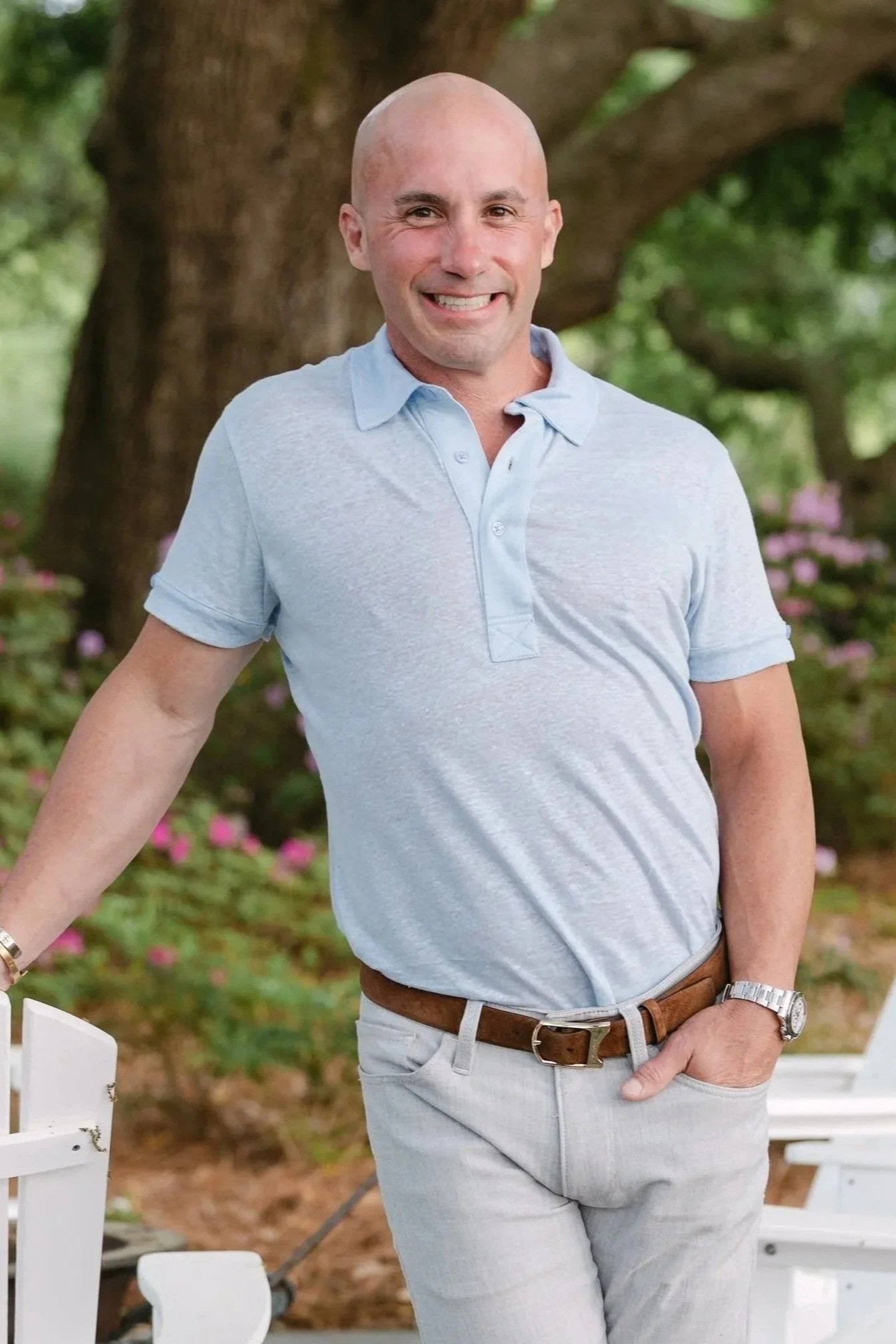 Dr. Matt Jamison with a shaved head smiling outdoors, wearing a light blue polo shirt and light-colored pants, standing in front of a tree and green foliage.