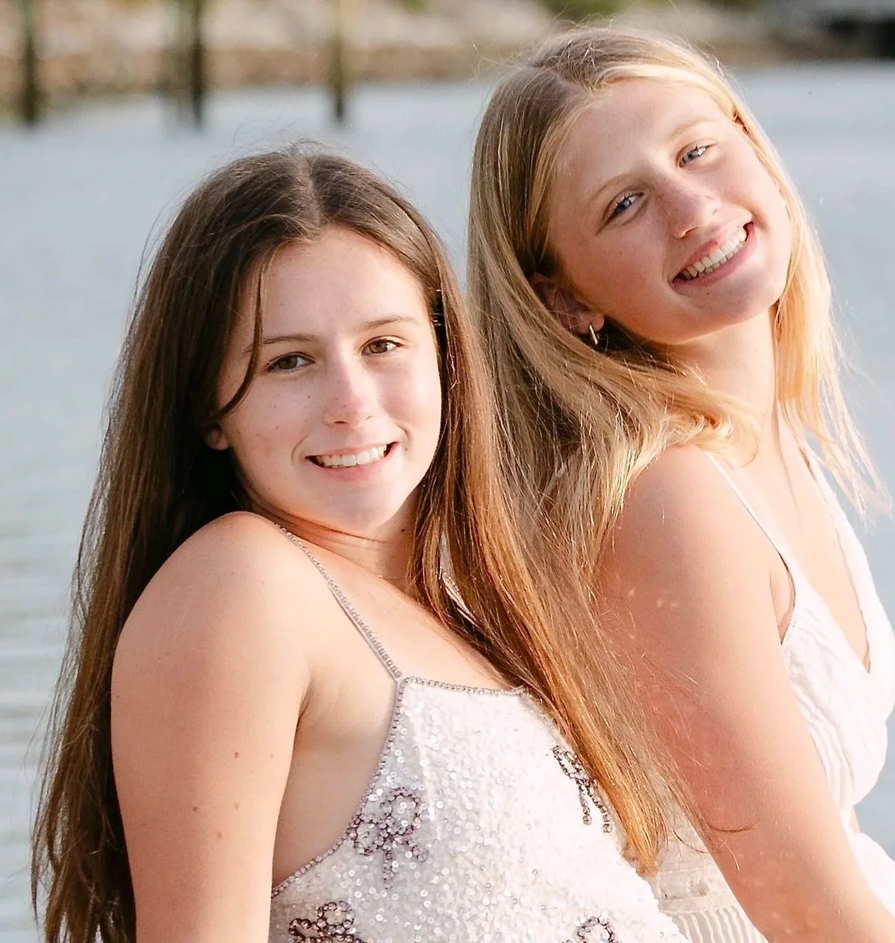 Two young women with long hair smiling, standing close together near a body of water with a fence in the background.