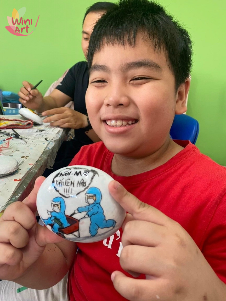 Boy in red shirt showing a decorated white rock featuring cartoon medical workers with a speech bubble, in a classroom or art studio with green walls.