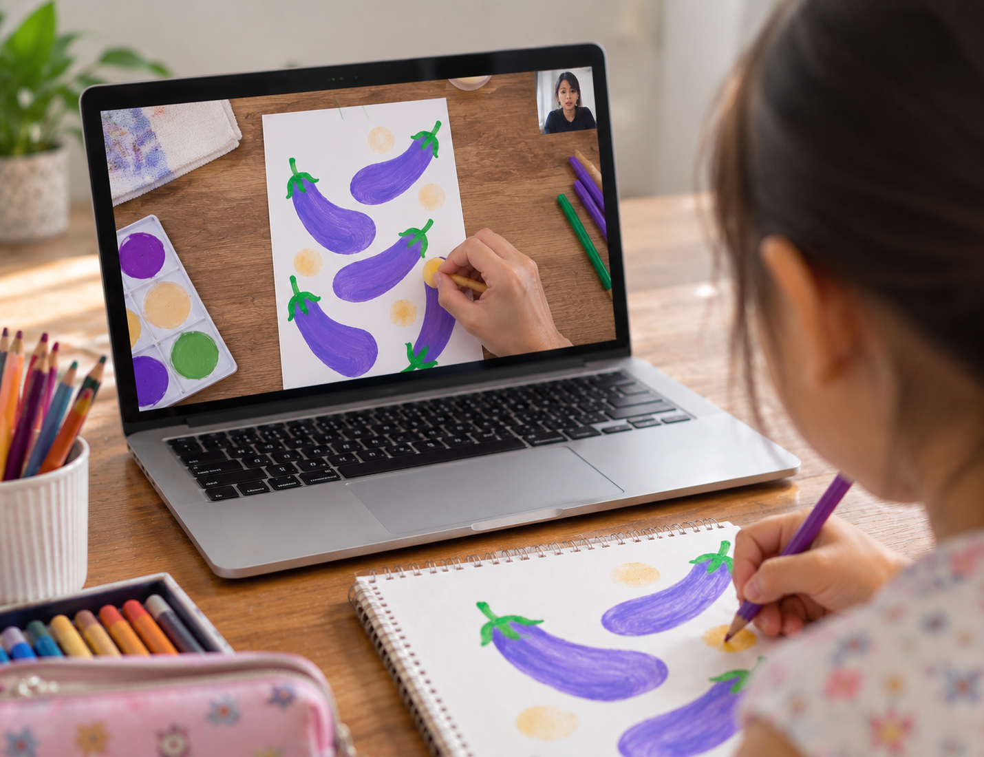 A girl is creating eggplant and dot watercolor paintings on paper during a video call on her laptop, which shows her artwork. Art supplies like colored markers, paints, and a sketchbook are on the wooden table.
