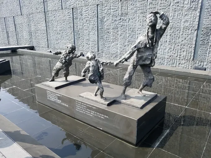 Bronze sculpture of a woman leading three children, pulling them along with a rope, on a rectangular base near a water feature against a textured wall.