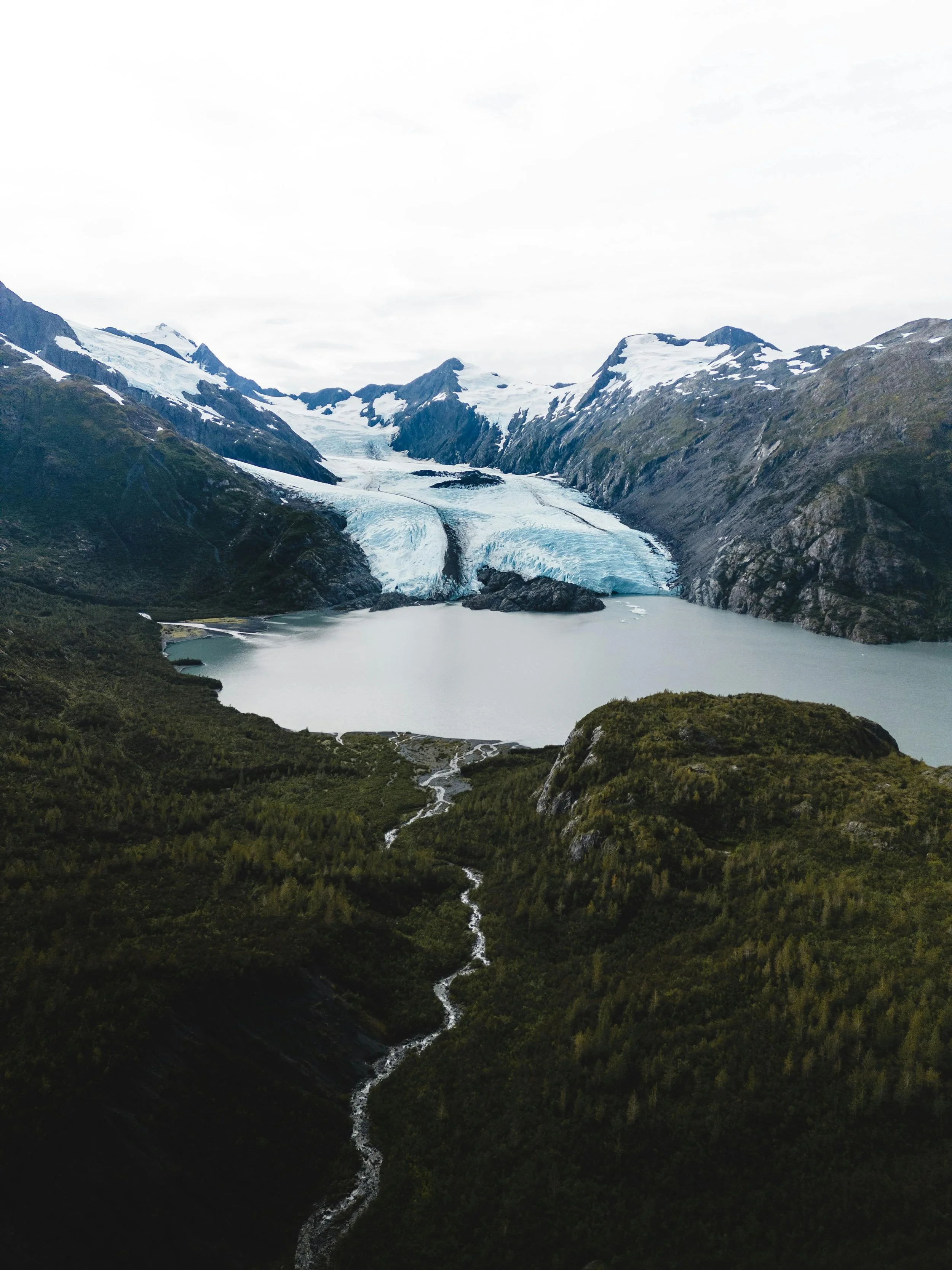 Scenic view of a glacier melting into a lake, surrounded by mountains with snow caps, lush green forest, and a winding river.