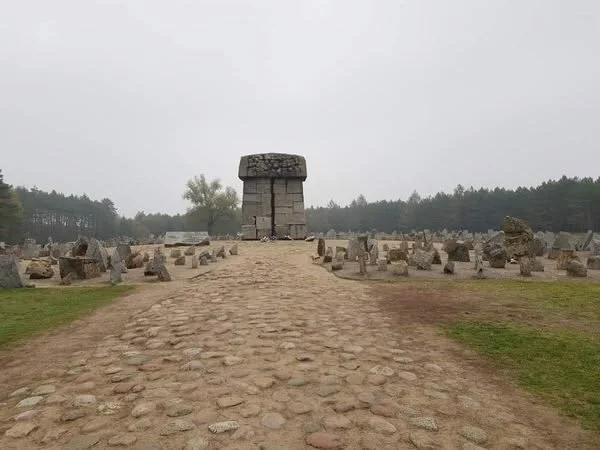 Ancient stone monument with a walkway leading to it, surrounded by smaller stone structures, under an overcast sky.