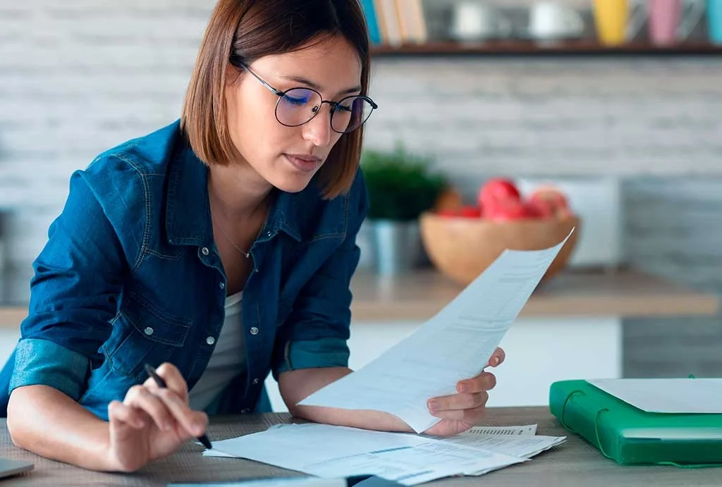 A woman with glasses and in a denim shirt reading and reviewing documents at a desk in a modern kitchen.