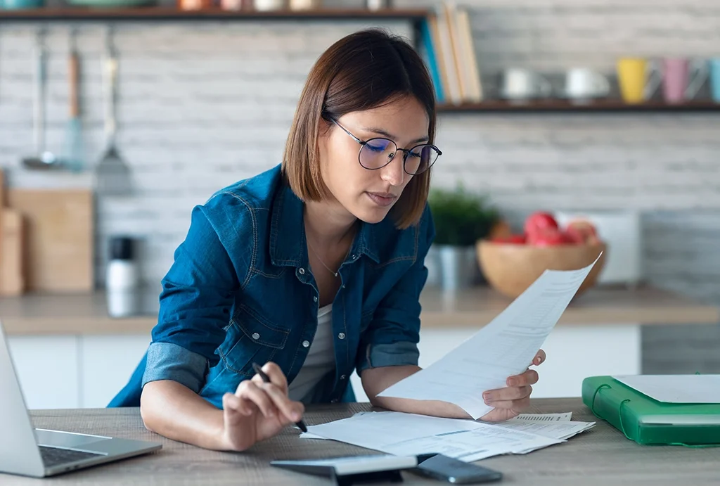 A woman with brown hair and glasses, wearing a denim shirt, looking at and holding a sheet of paper while seated at a desk in a modern kitchen.