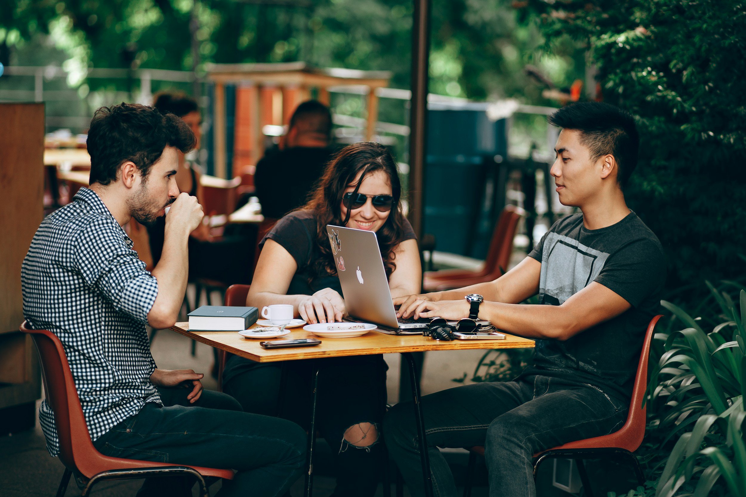 Three young adults sitting at a table outdoors, working on a laptop, with drinks and notebooks, surrounded by trees.
