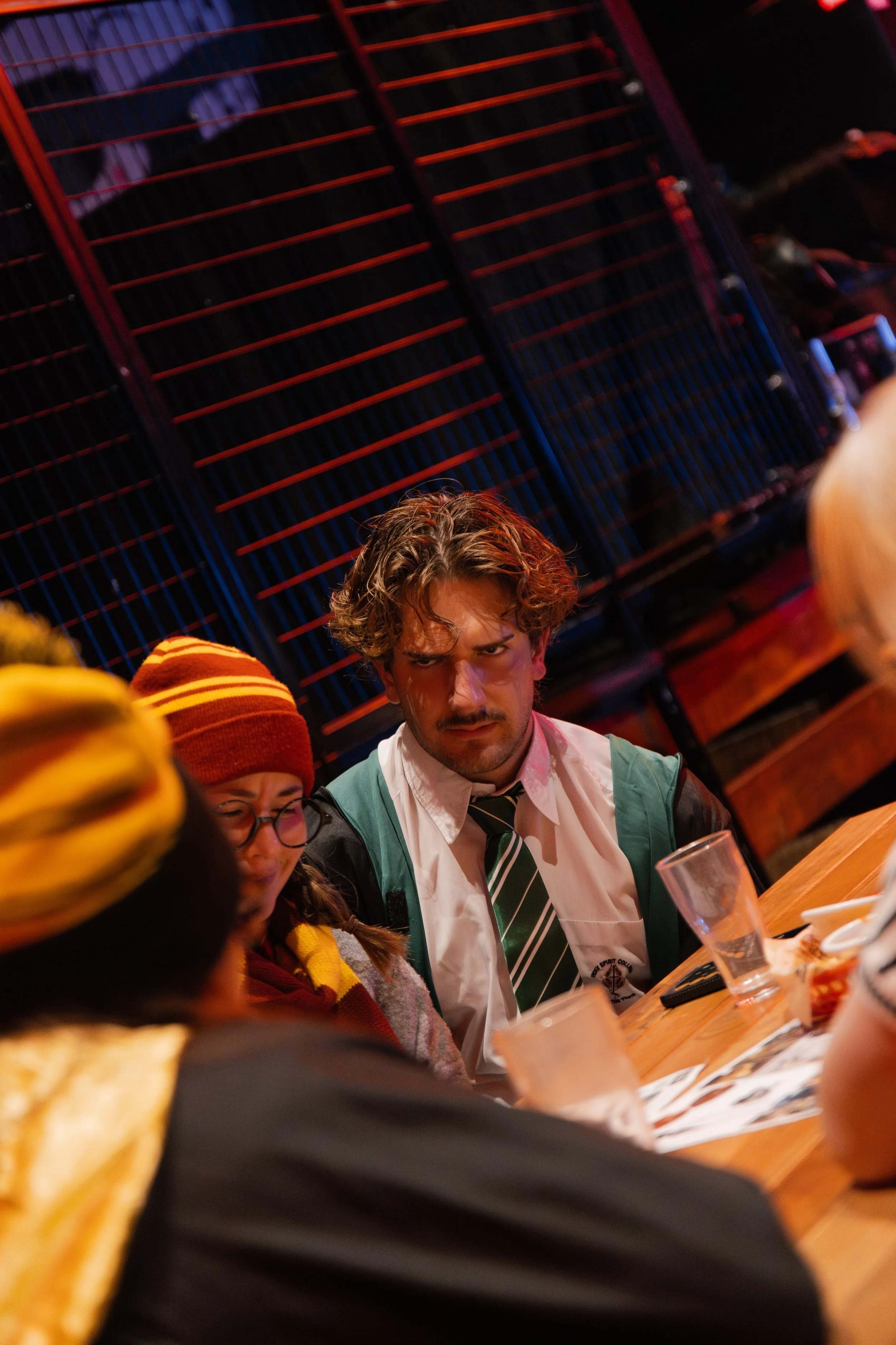 People sitting at a restaurant table, with food and drinks, in a dimly lit setting with a large red and black metal wall in the background.
