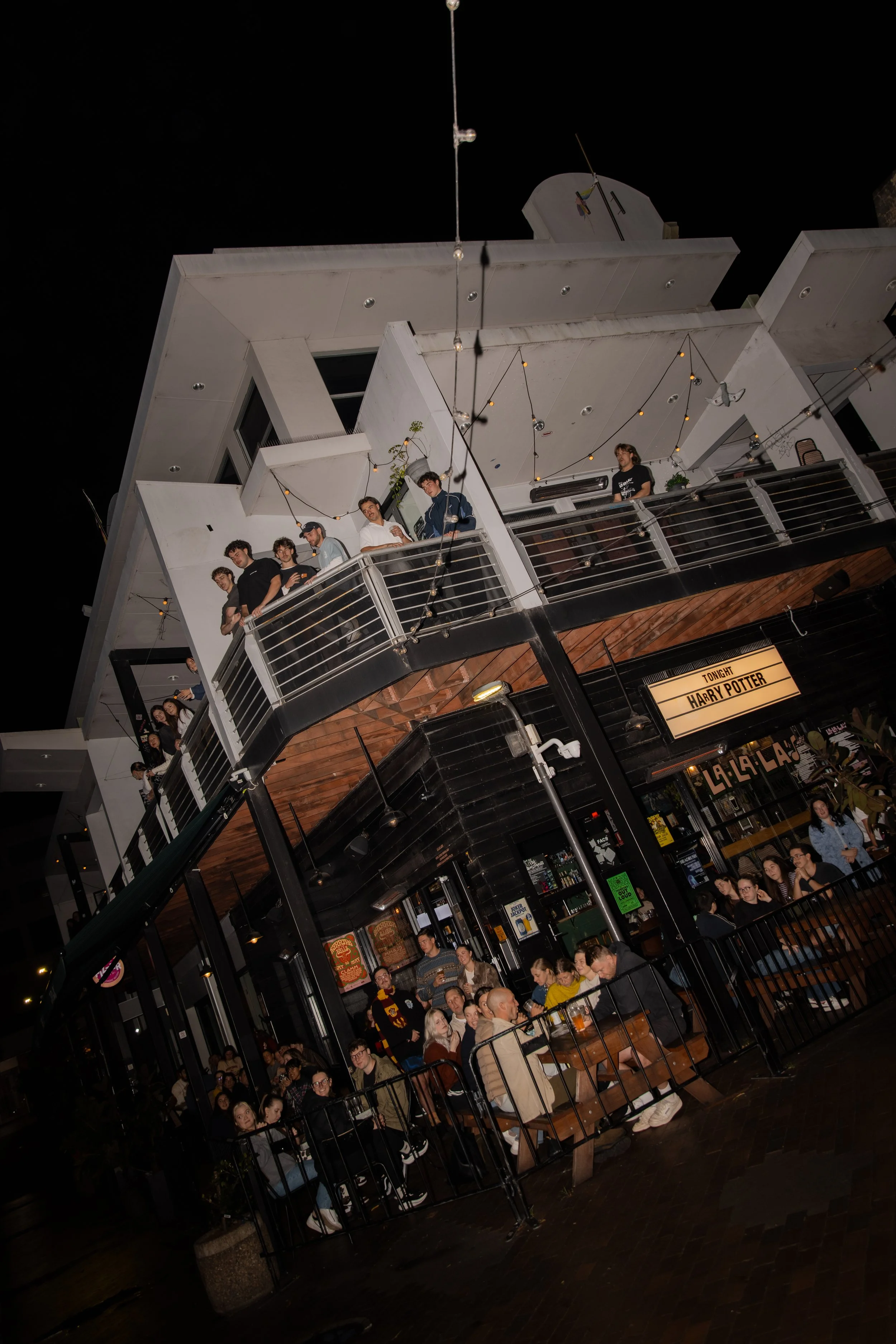 Nighttime scene of patrons in a multi-level bar or restaurant with modern decor, string lights, and a sign indicating a Harry Potter movie screening.