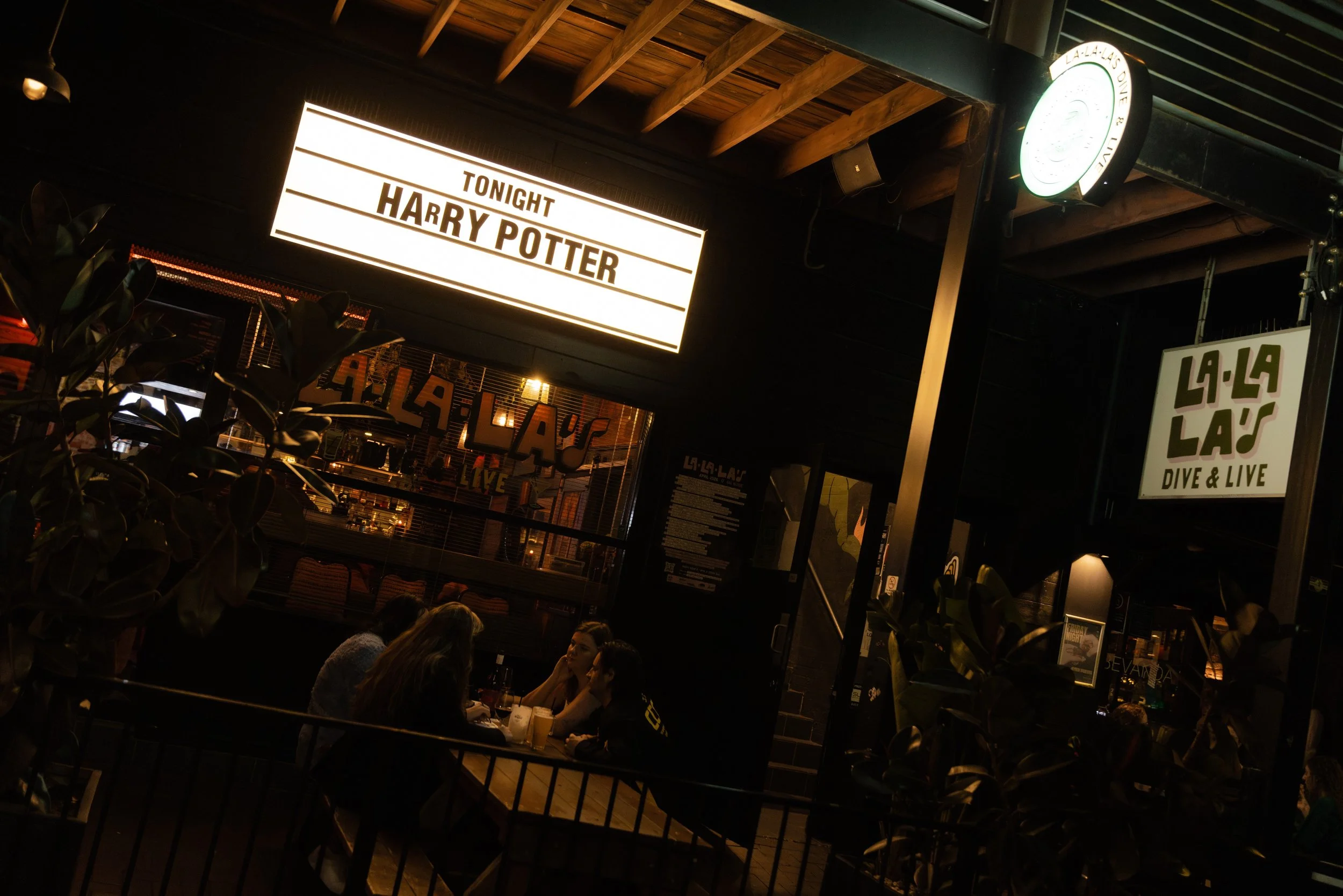 Interior of a bar or restaurant at night with people socializing. A brightly lit sign advertises a Harry Potter-themed event tonight. The space features wooden ceiling beams and plants near the entry.