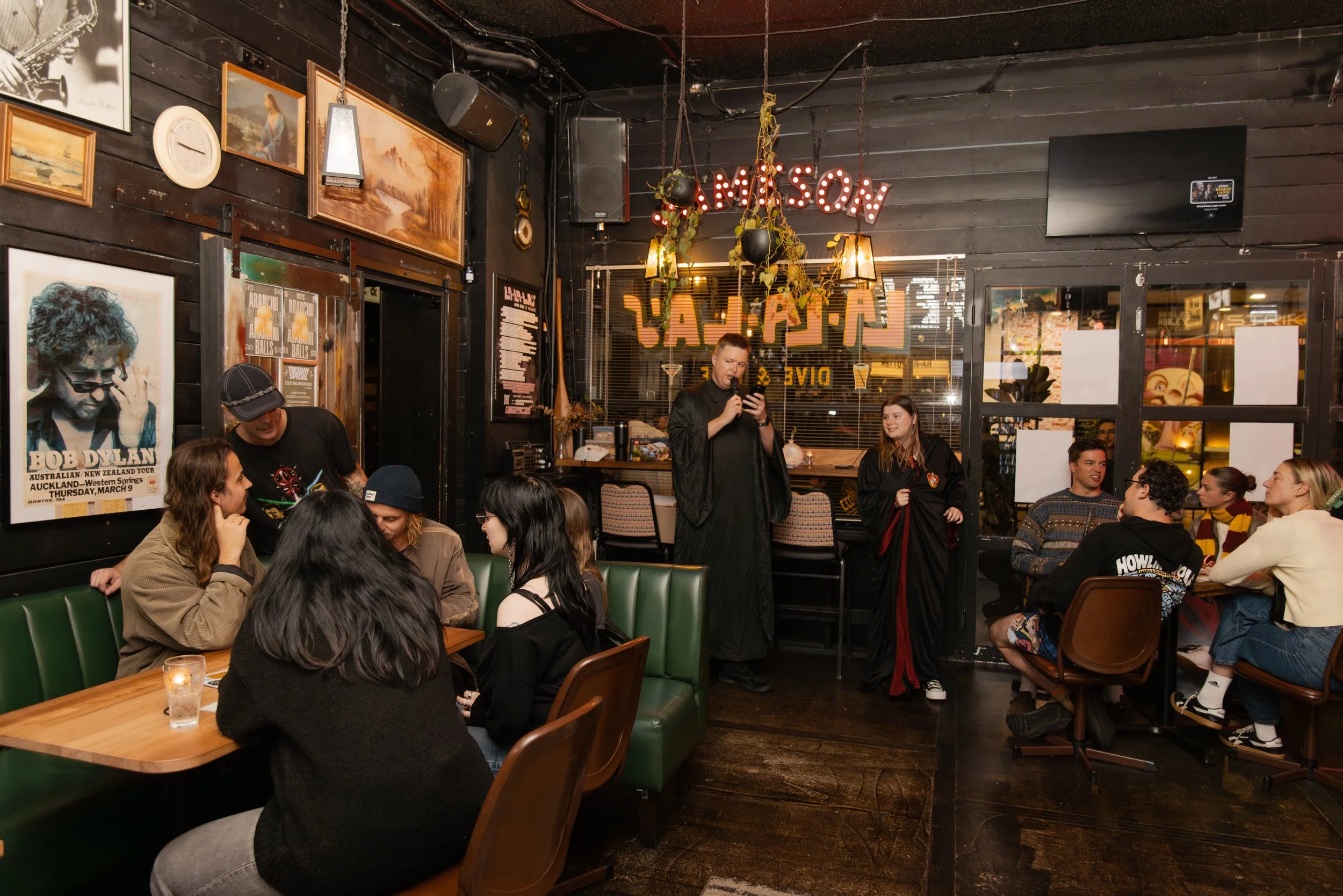 A group of people sitting around tables in a dimly lit restaurant or bar, with a performer standing in the background holding a microphone. The wall is decorated with framed pictures and artwork, including a poster of Bob Dylan, and illuminated by wa