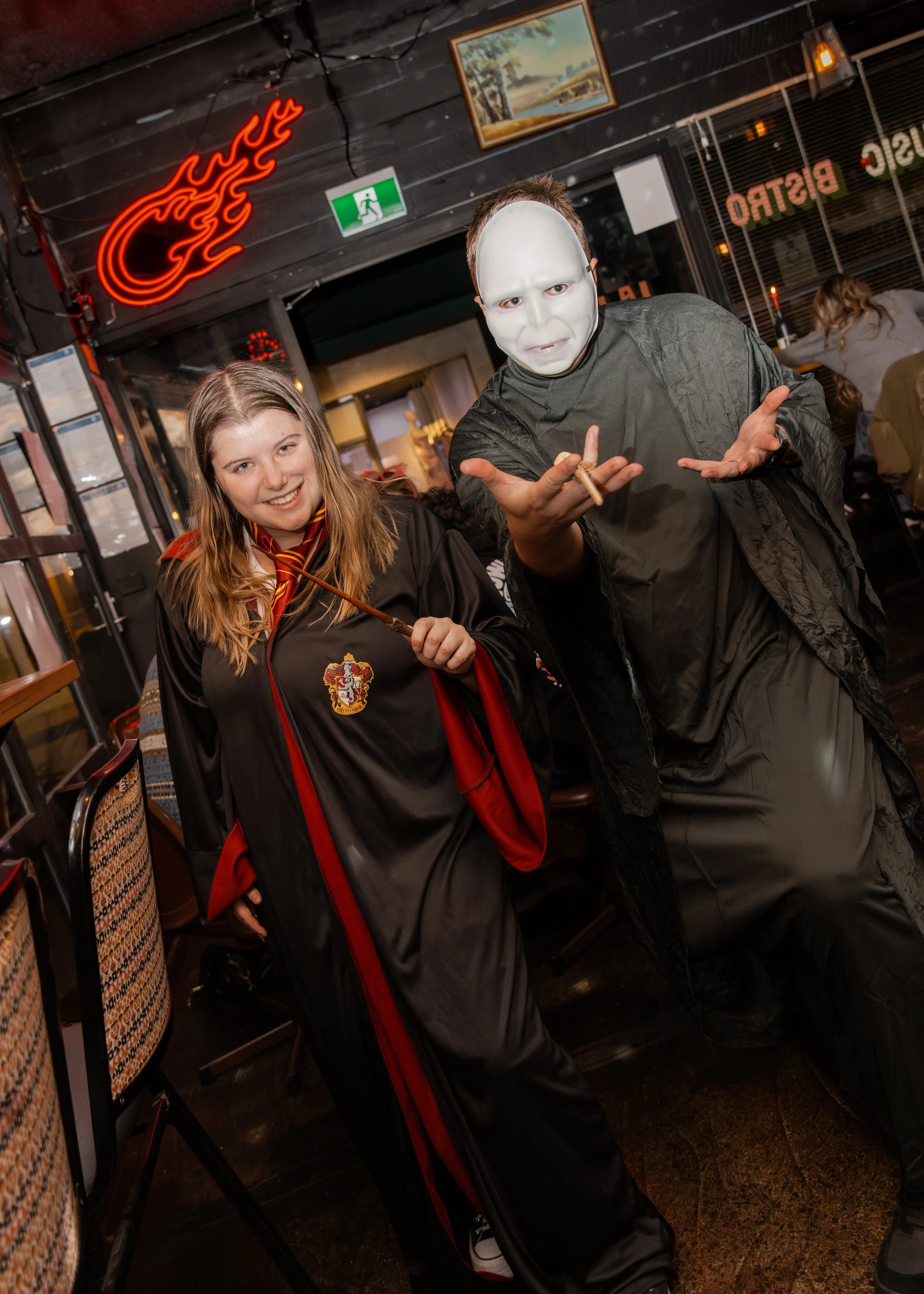 Person dressed as Michael Myers from Halloween, wearing a white mask and dark clothing, in a restaurant with a woman in a black and red robe smiling next to them.