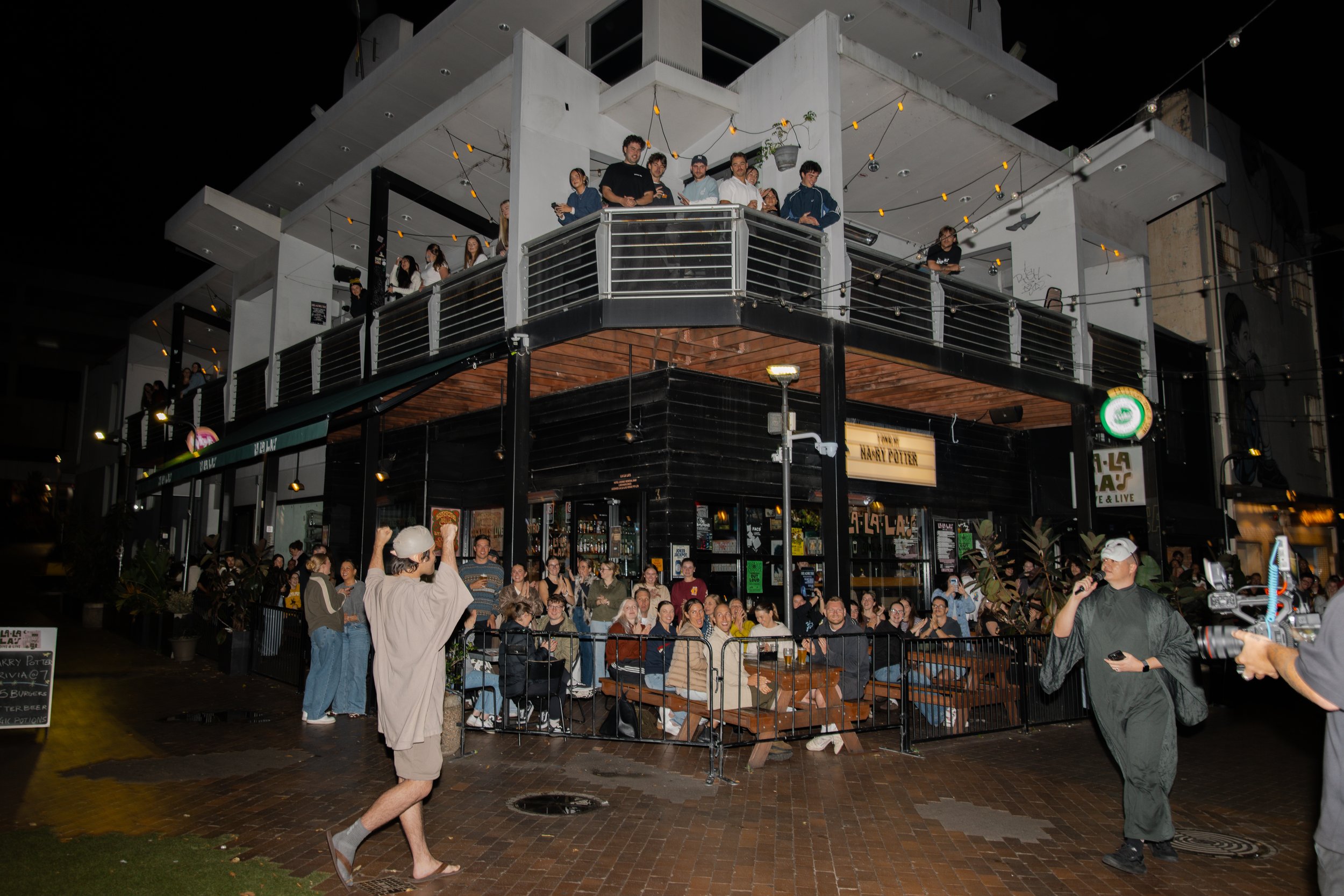 A crowd of people gathered outside a two-story building at night, with some on the upper balcony and some on the lower level, watching a performer with a microphone. The building has string lights and a sign that says "LA LA LA" on the corner. The au