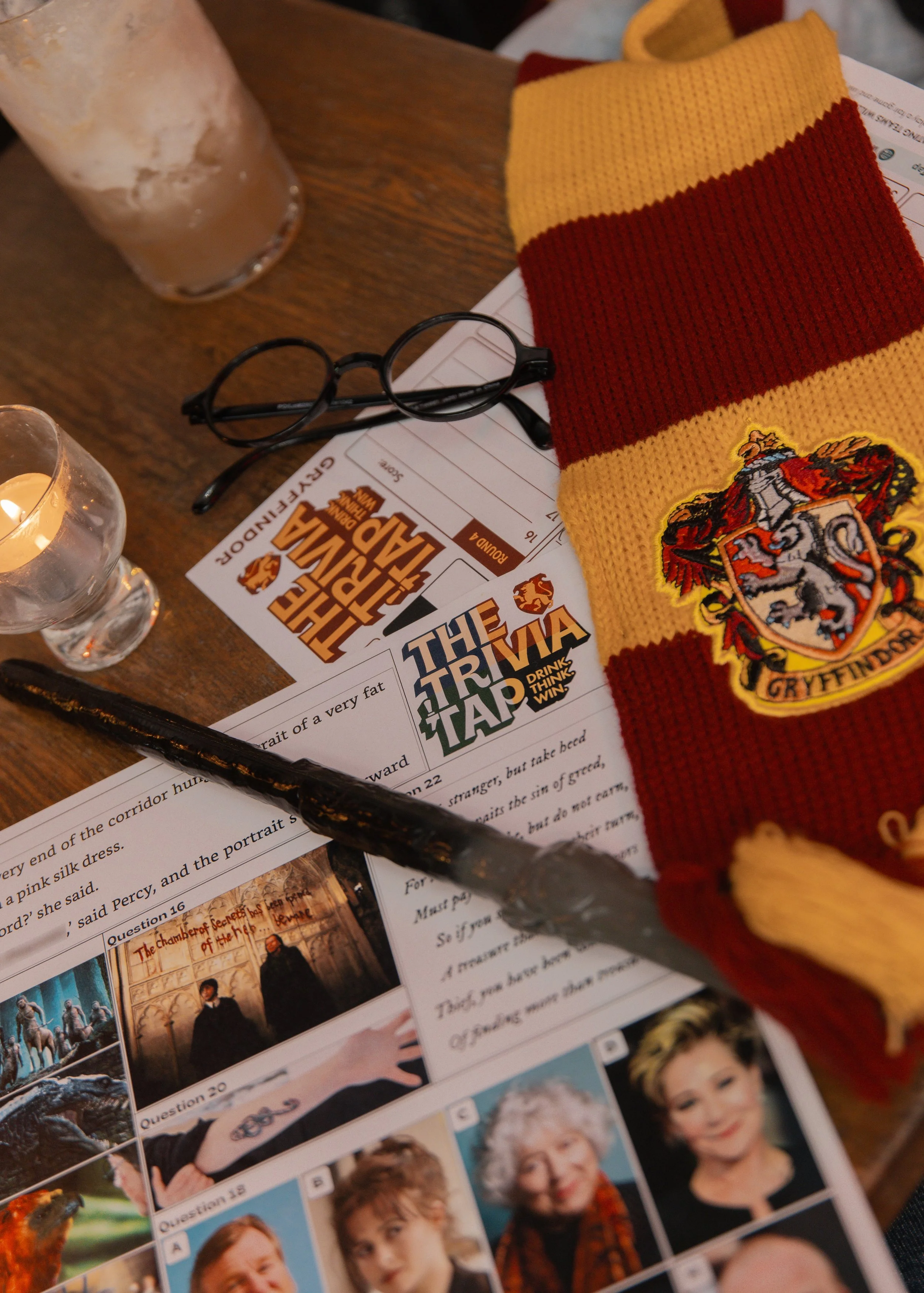 Close-up of a Harry Potter themed quiz or trivia game on a table, with a red and yellow Gryffindor scarf, reading glasses, a candle, a tall glass of iced beverage, and quiz sheets with photos of children and Harry Potter characters.