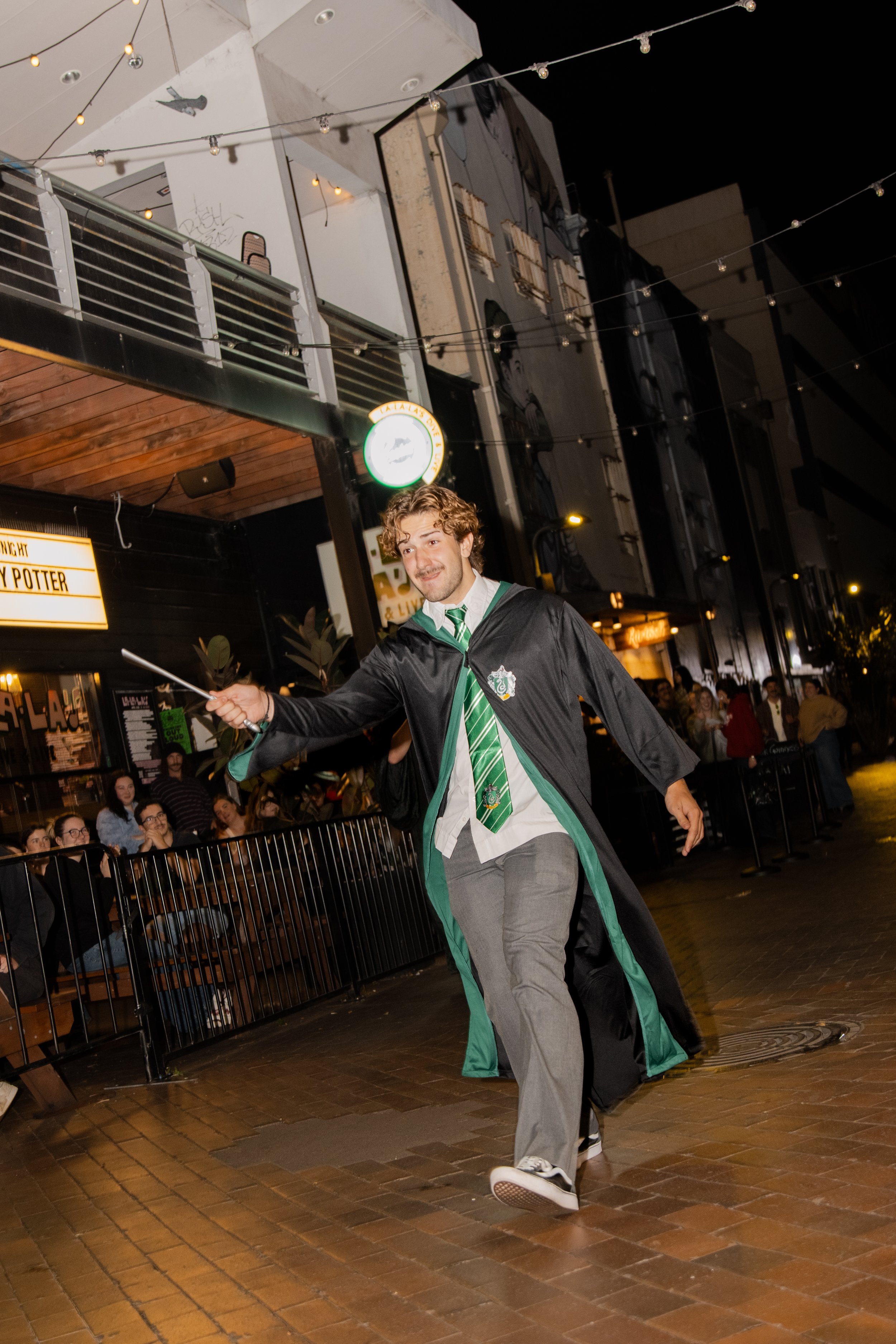 A young man dressed in Gryffindor robes from Harry Potter holding a wand and smiling, walking outdoors at night with festival lights and a crowd in the background.