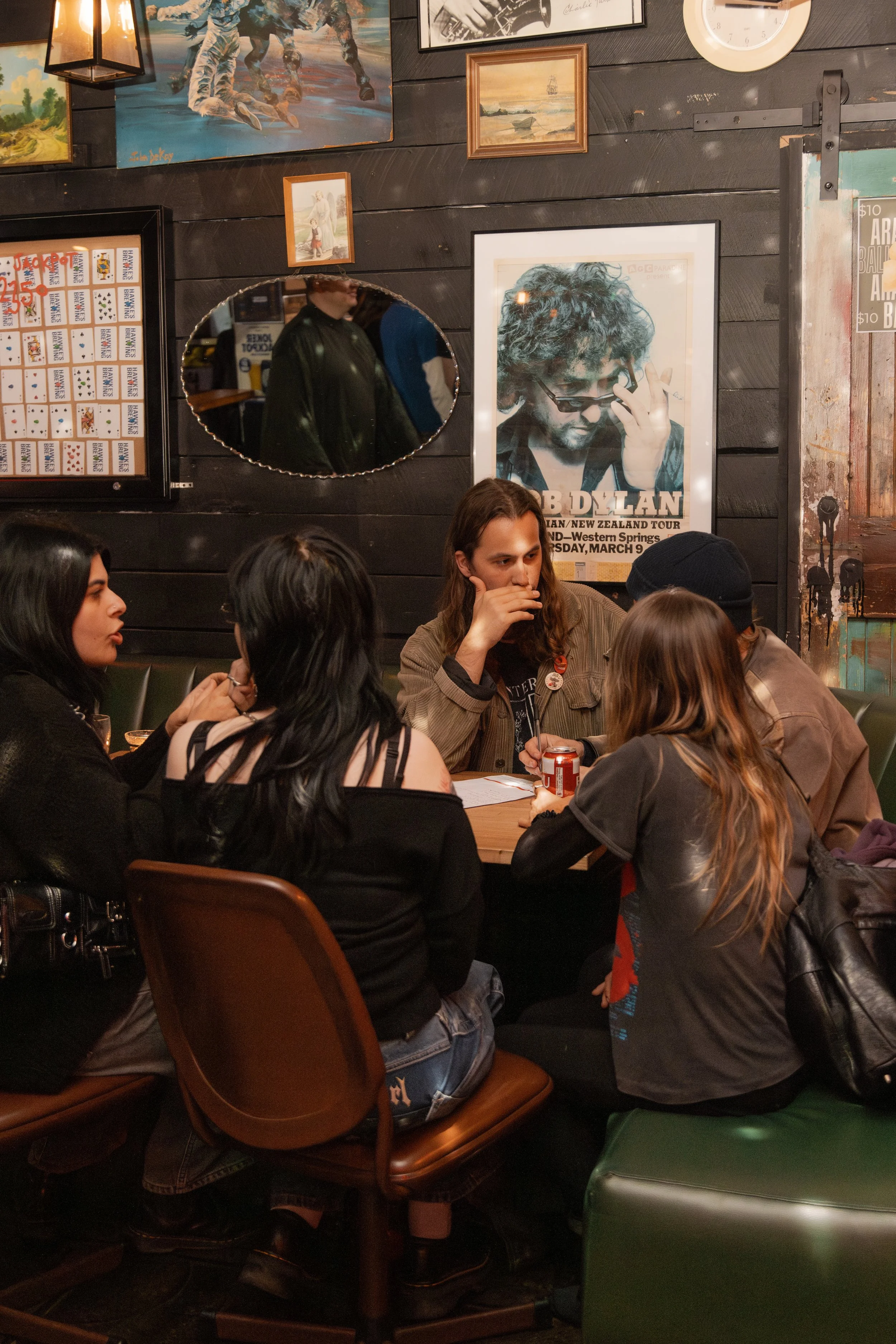 A group of people sitting around a table in a restaurant with dark walls decorated with paintings and a poster of Bob Dylan. Some of them are talking, and one is holding a can. There is a small mirrored surface reflecting part of the scene.